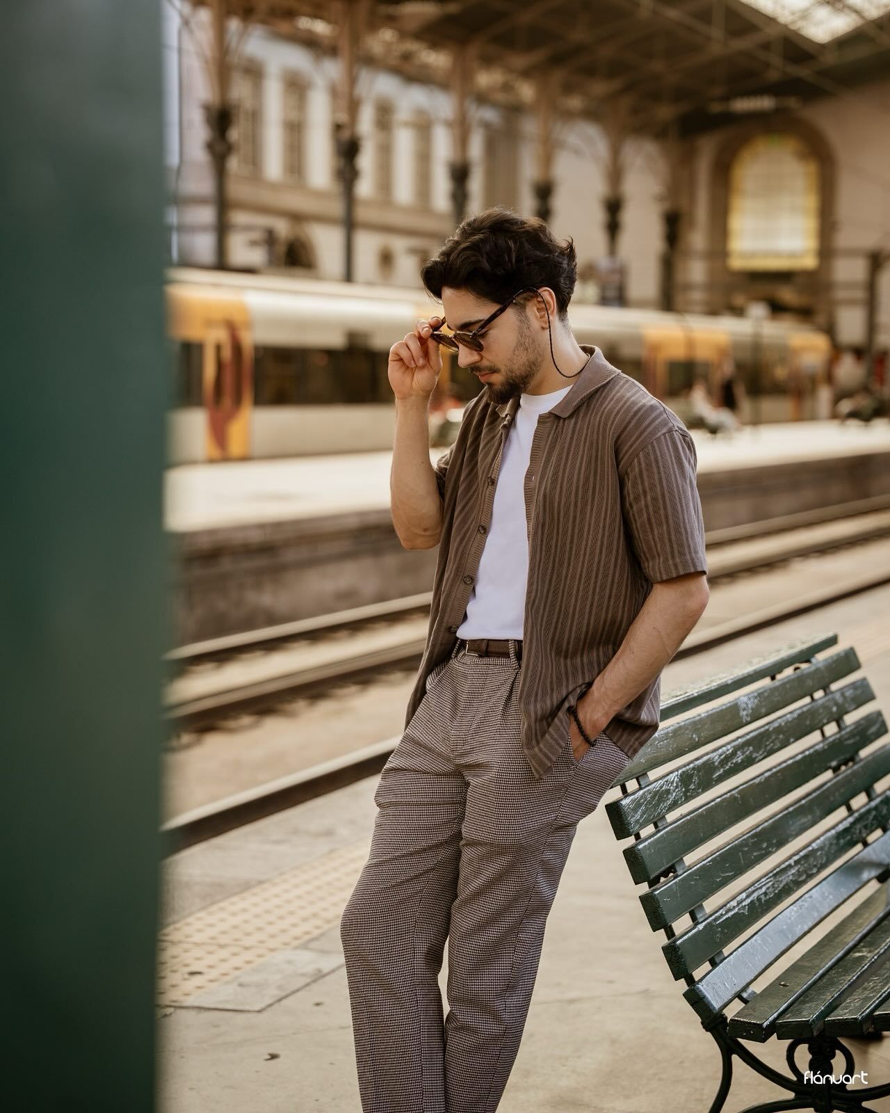 Stylish man in striped brown shirt and trousers adjusting sunglasses on a train station platform.