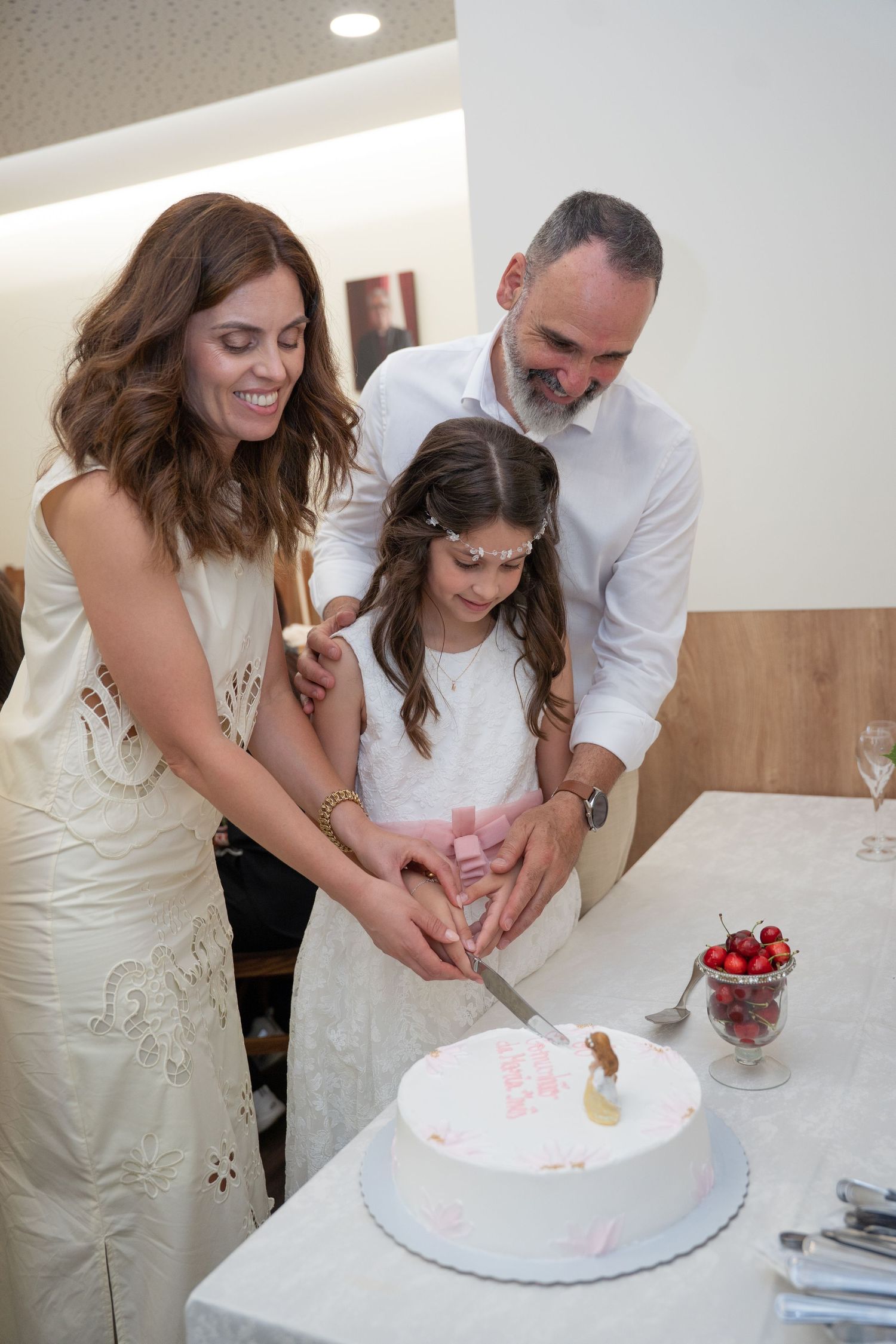 Family cutting a white cake with pink decorations together at a celebration, with cherries on the table nearby.