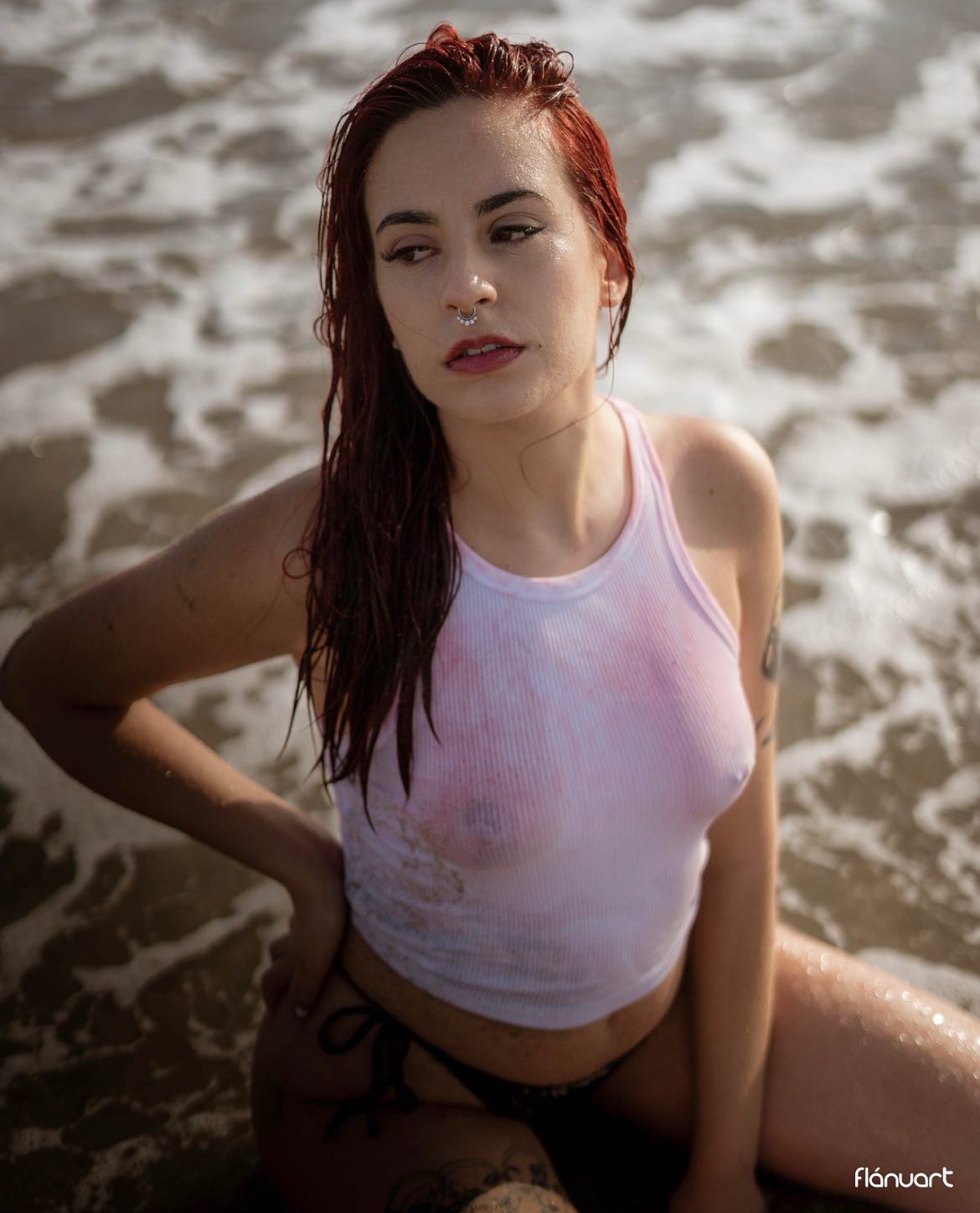Red-haired woman in wet white tank top sitting in ocean waves on sandy beach.