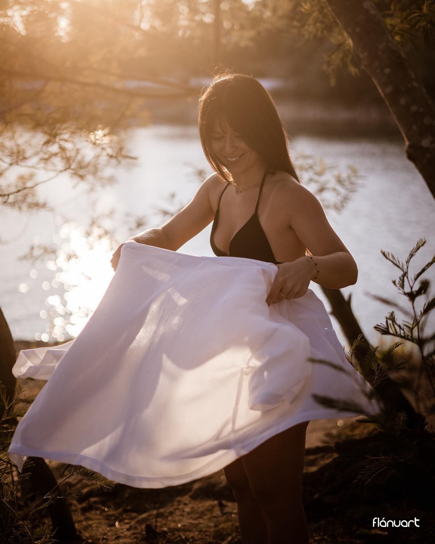 Woman in black swimsuit twirling white skirt near sunlit river surrounded by trees.