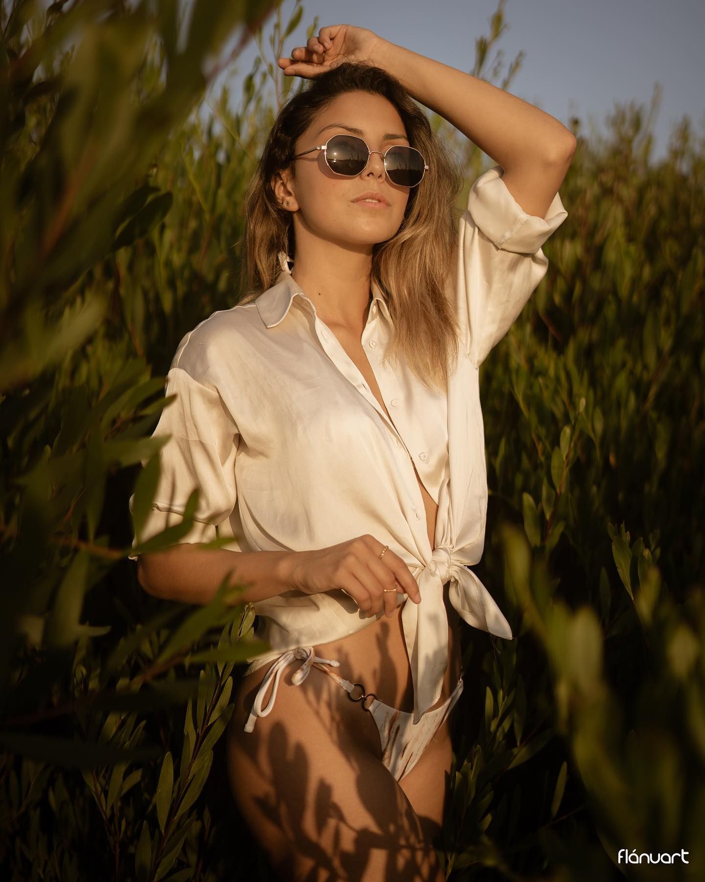 Young woman in a tied white shirt and sunglasses posing in a lush green field at golden hour.