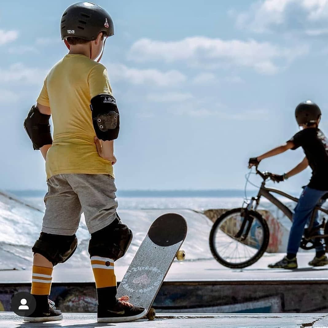 Young boy in yellow shirt with skateboard and protective gear watches another child ride a bike at a skatepark.