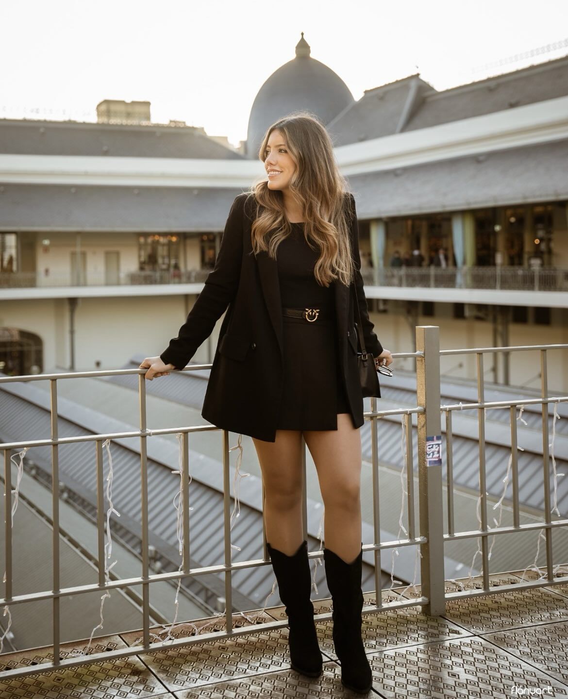 Stylish woman in all-black outfit with knee-high boots posing on rooftop terrace at sunset.