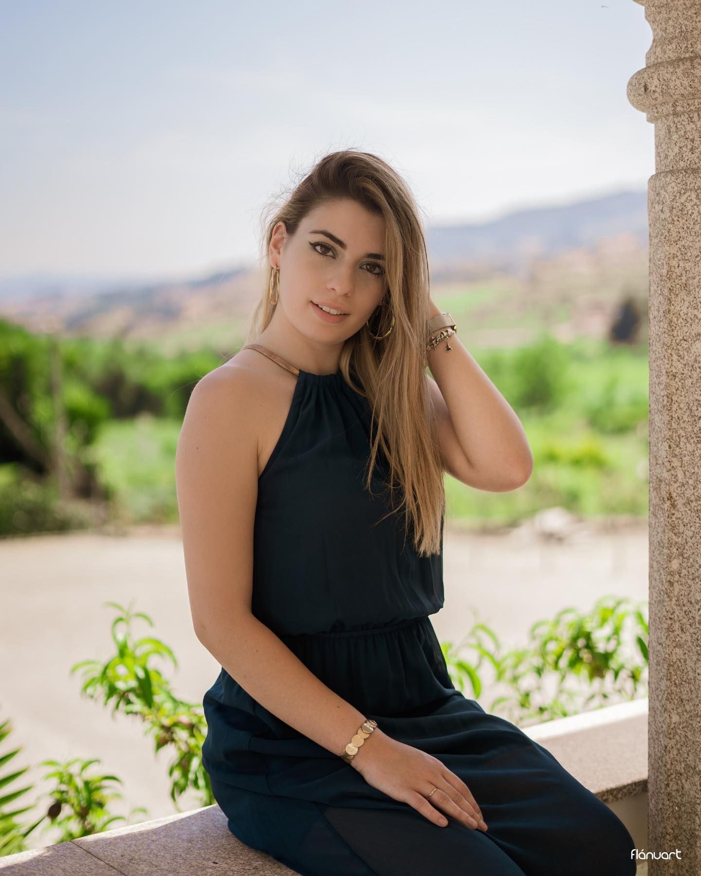 Blonde woman in dark halter jumpsuit sits on stone ledge with green landscape behind her.