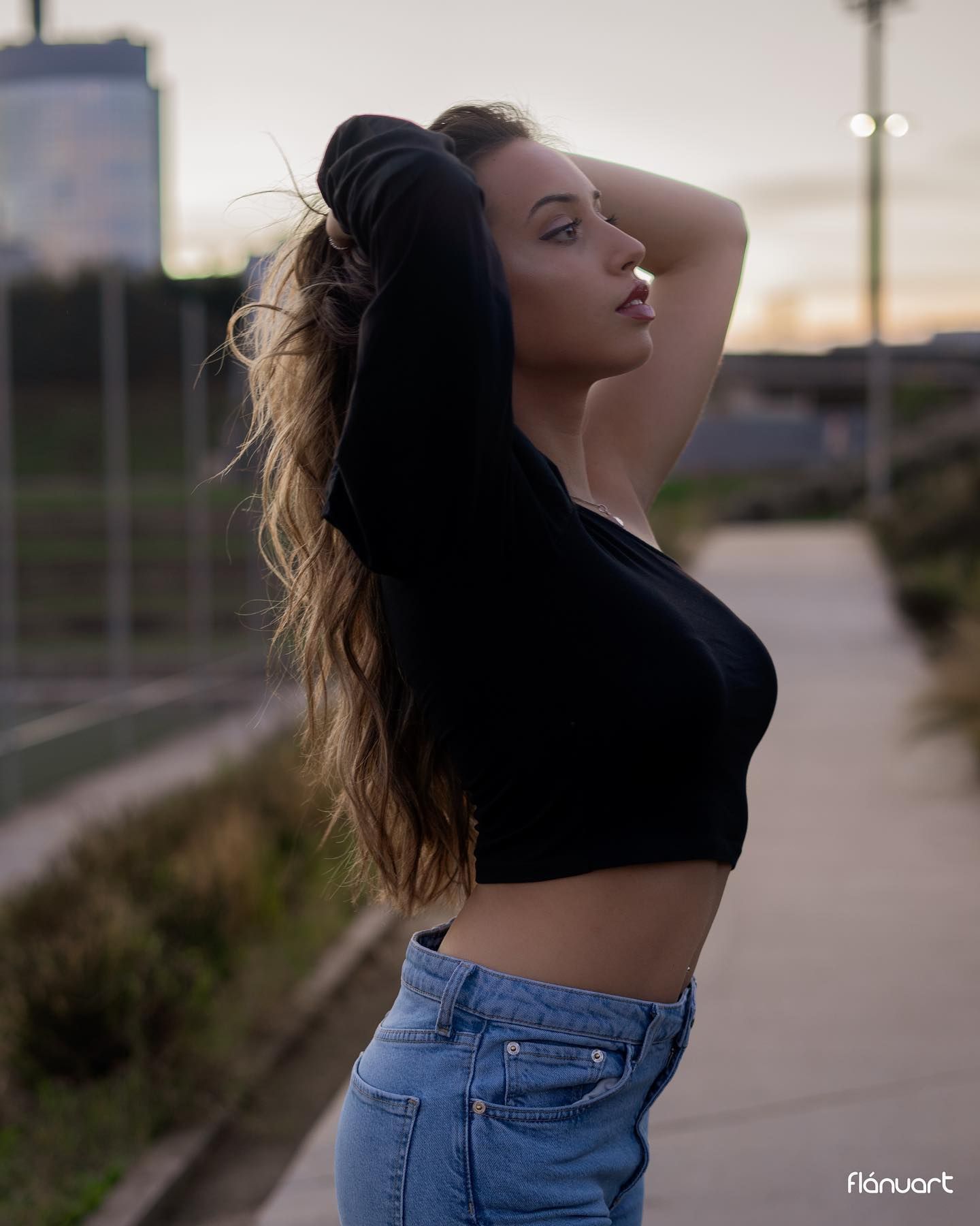Woman in black crop top and jeans posing outdoors at dusk with hair swept back.
