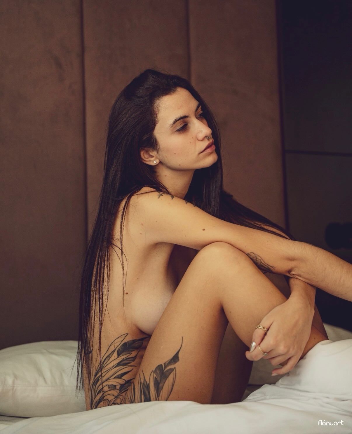 Brunette woman with leg tattoo sitting on white bed in moody hotel room lighting.