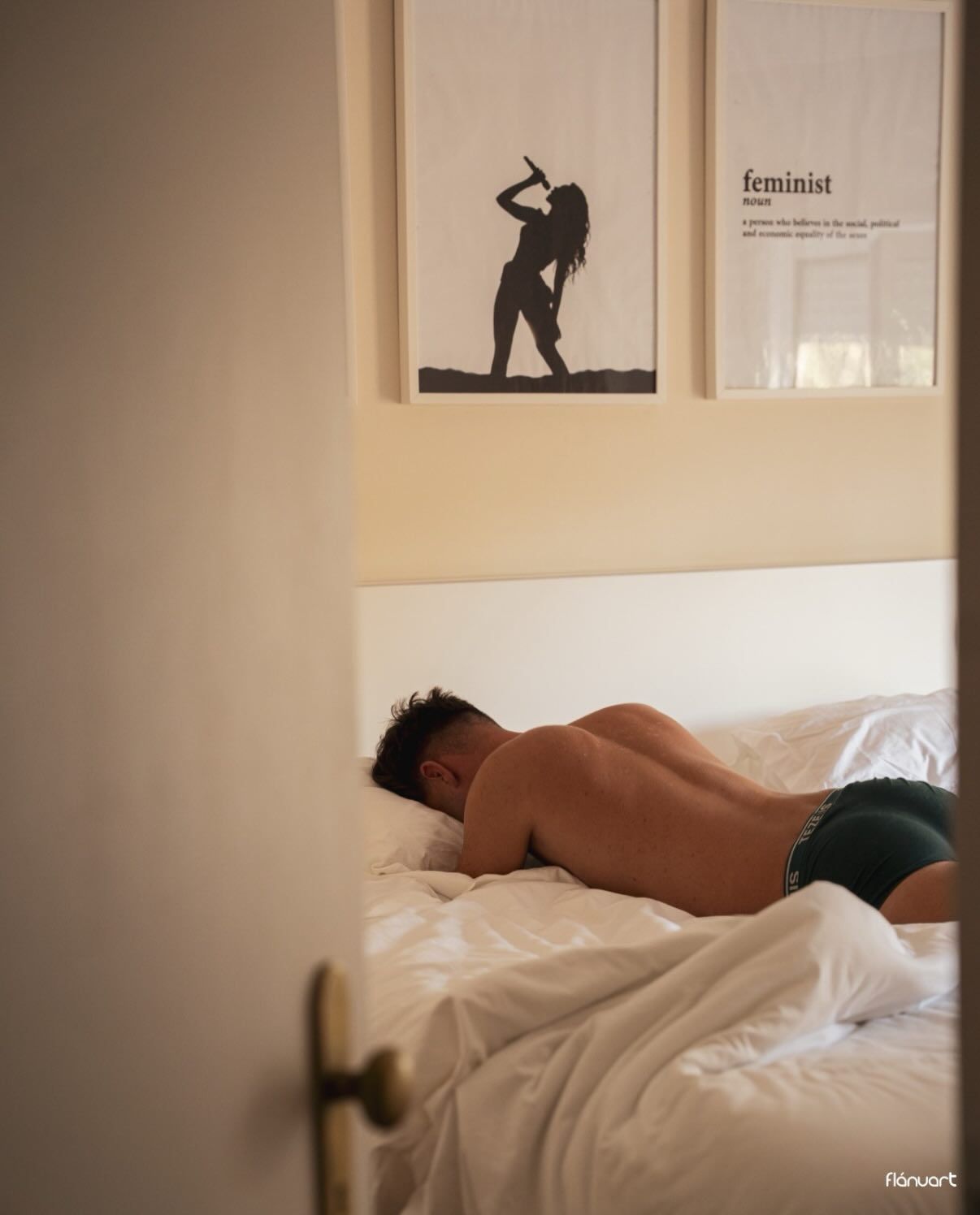 Shirtless man lying face down on white bedding near framed feminist wall art in a bright bedroom.