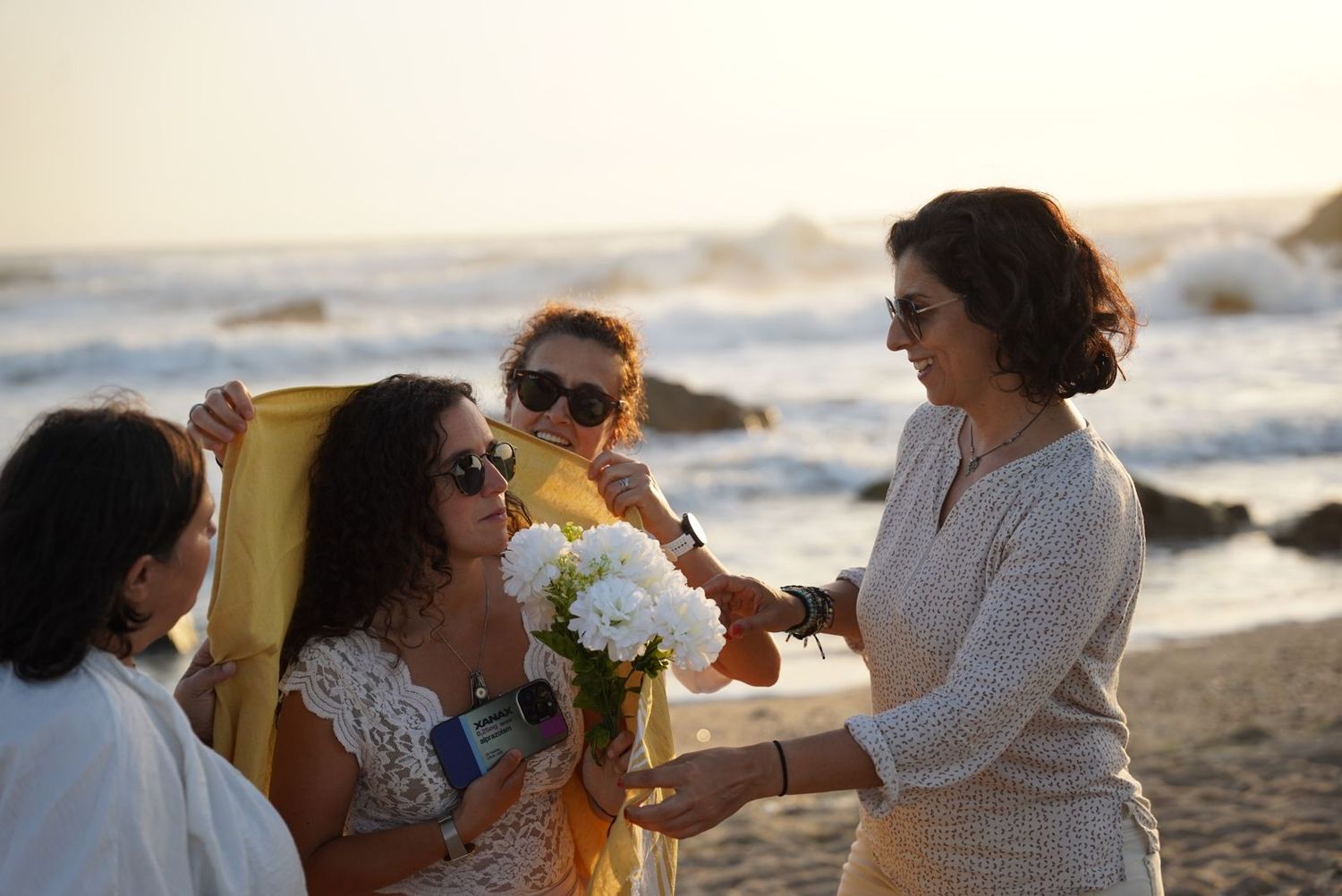 Women celebrating on a beach at sunset, one holding white hydrangeas, with golden light behind them.