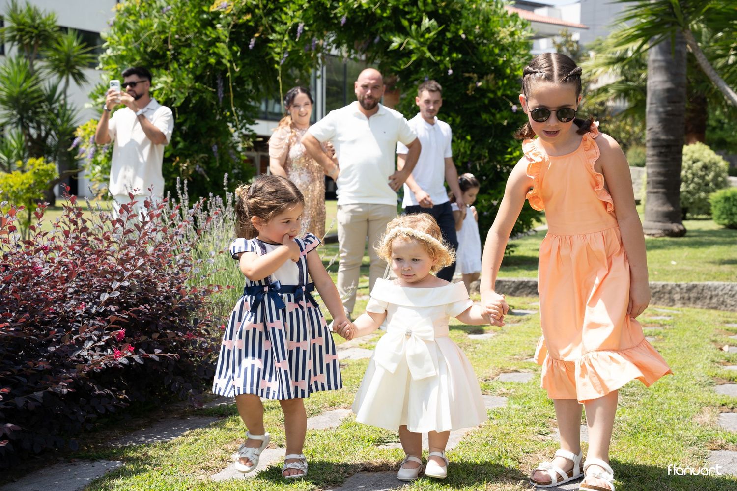 Three young girls in elegant dresses walk hand in hand in a sunny garden during a special occasion.