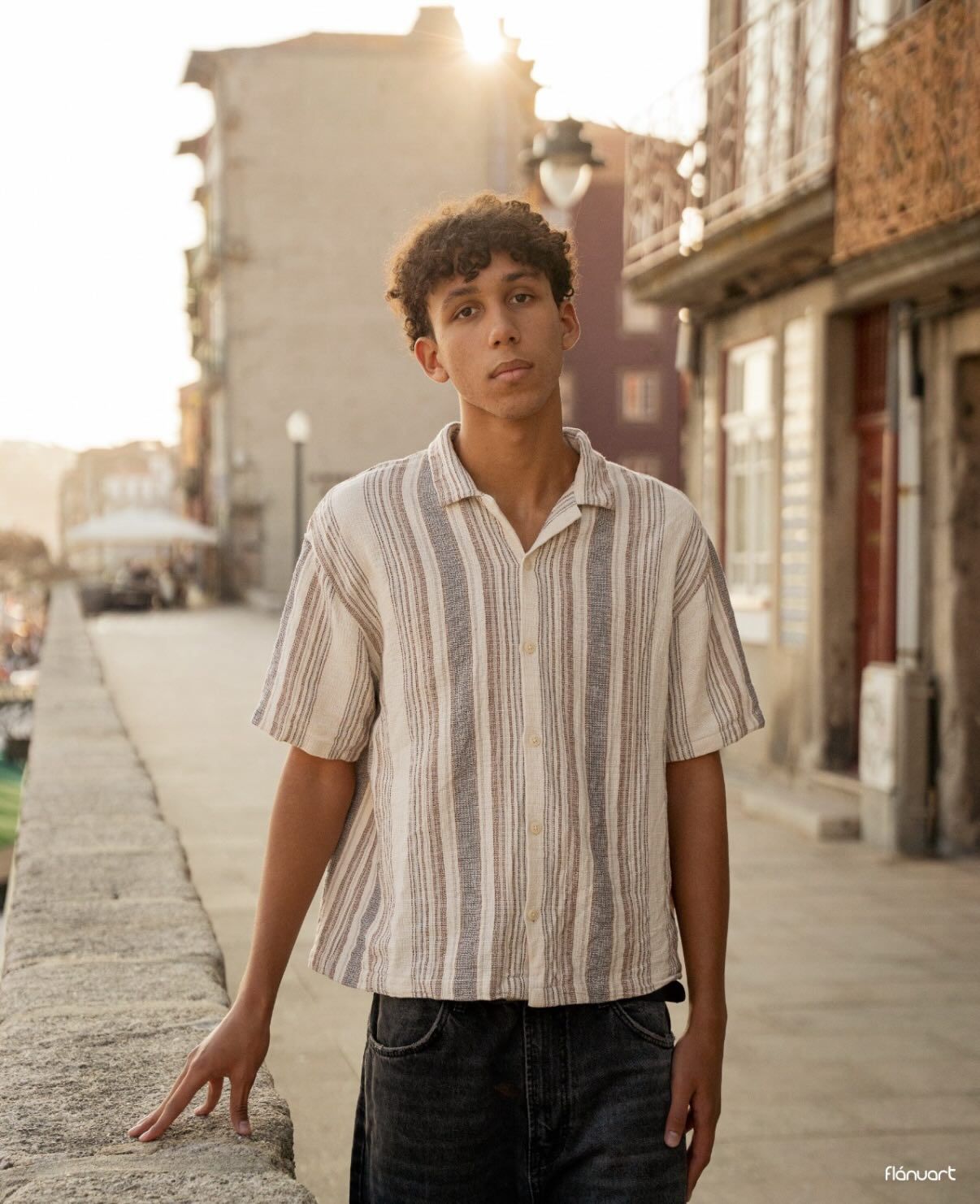 Young man in striped linen short-sleeve shirt standing on a cobblestone street at golden hour.