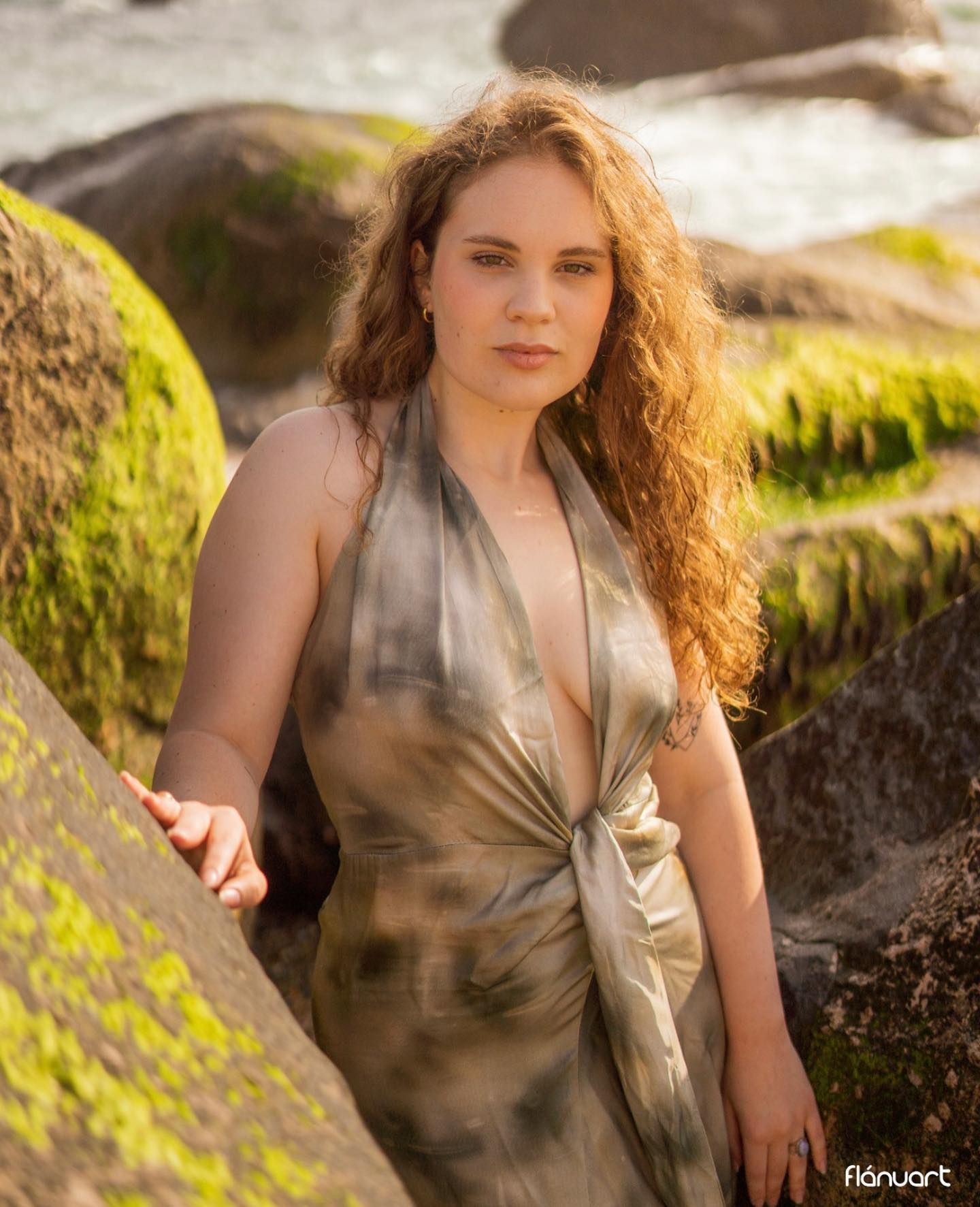 Young woman with curly hair in silver halter dress posing among mossy coastal rocks.