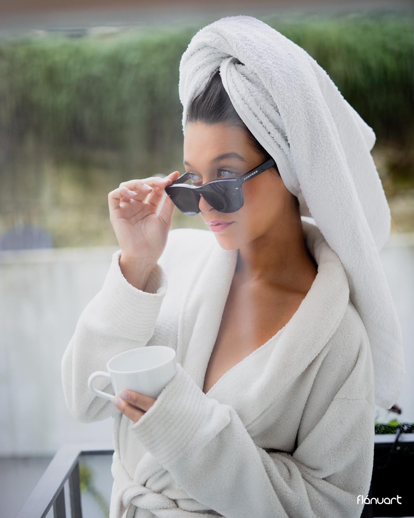 Elegant woman in white bathrobe and towel wrap wearing sunglasses holding a coffee cup outdoors.