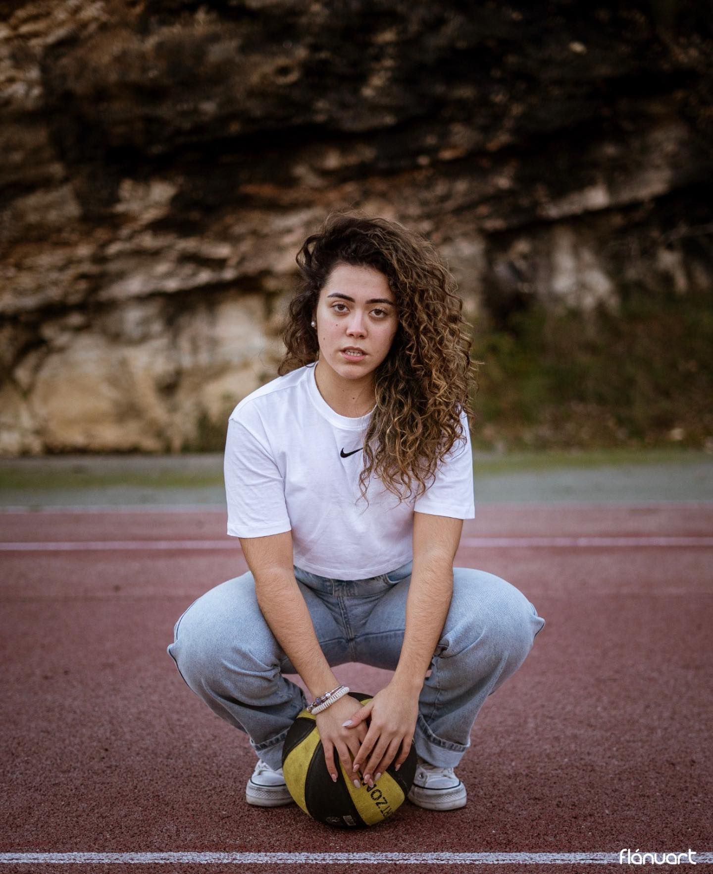 Young woman with curly hair crouching with a basketball on an outdoor sports court.