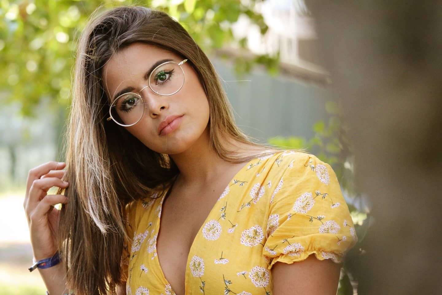 Young woman in a yellow floral dress and round glasses posing near a tree in a park.