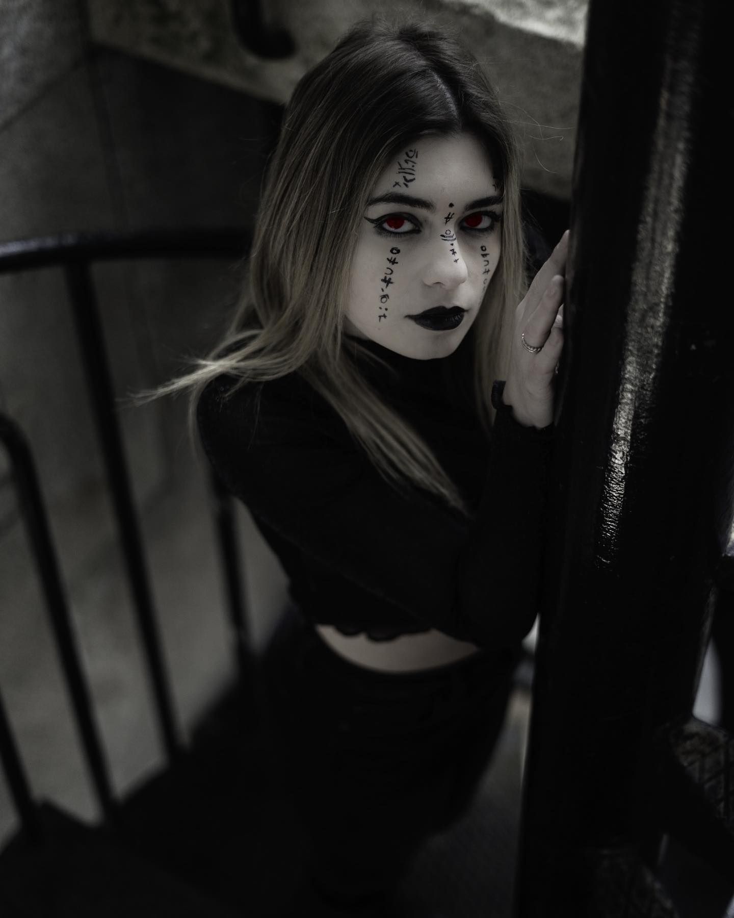 Woman with dramatic gothic face paint and dark makeup posing on a staircase in black outfit.