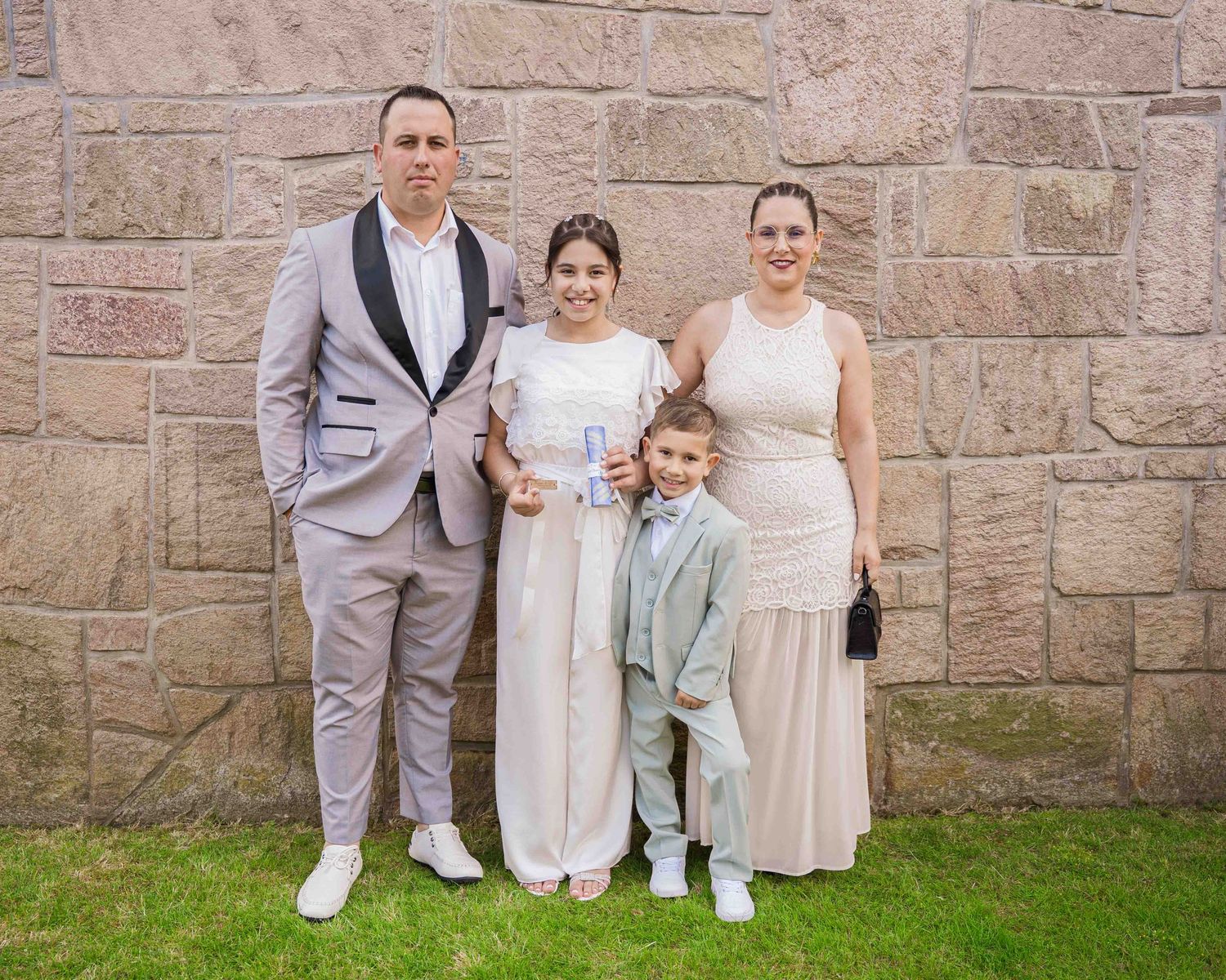A family of four poses in formal attire against a stone wall for a First Communion event.
