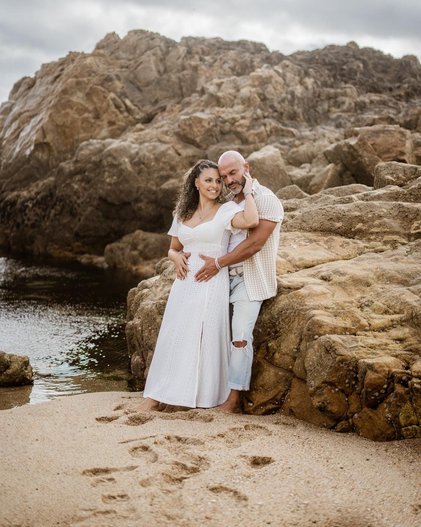 A smiling couple embraces against large rocks on a sandy beach with footprints in the sand.