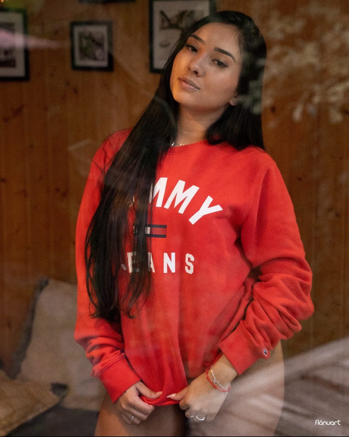 Young woman with long dark hair wearing red Tommy Jeans sweatshirt posing indoors.