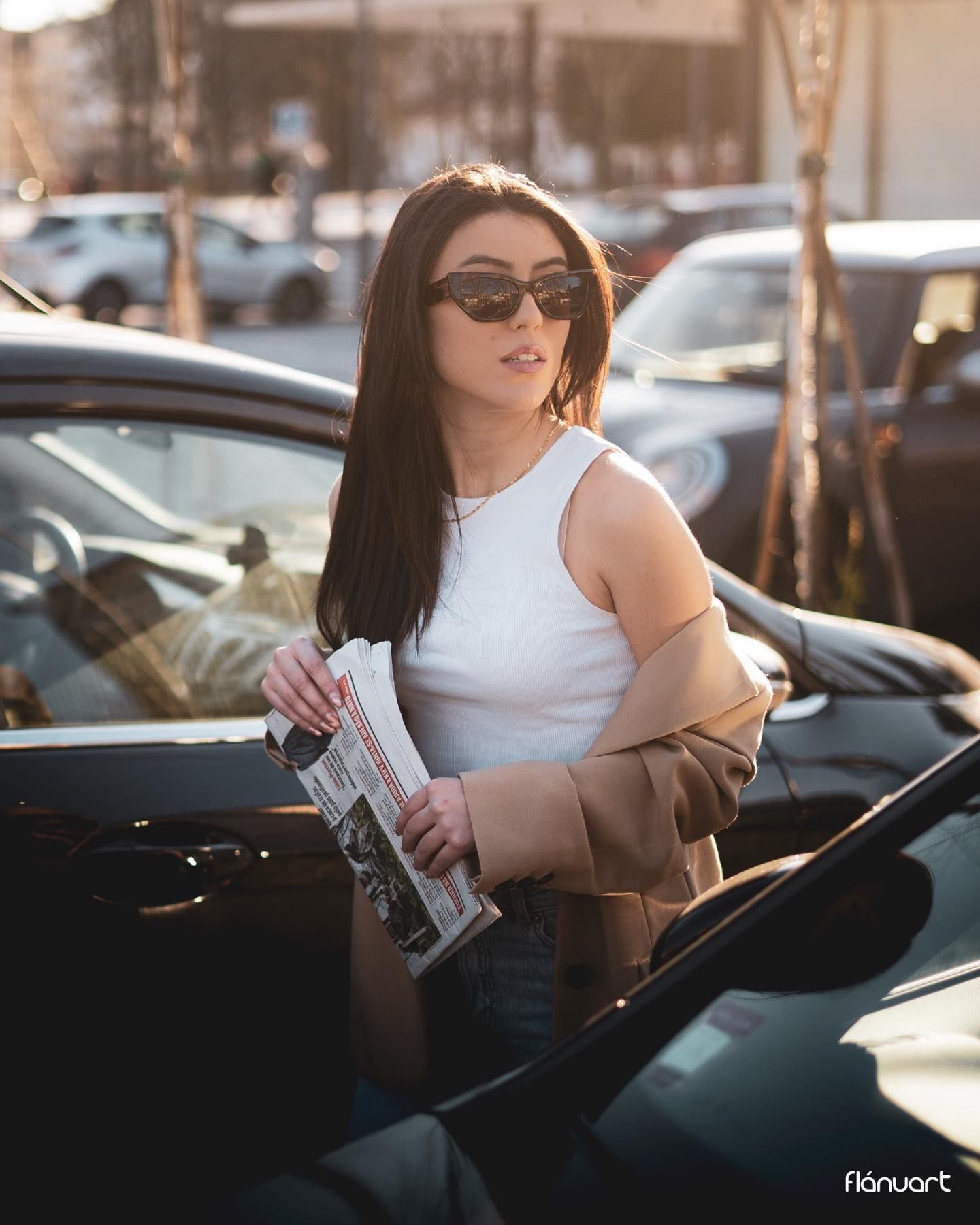 Stylish woman in white tank and beige blazer holding magazine beside a black car at sunset.