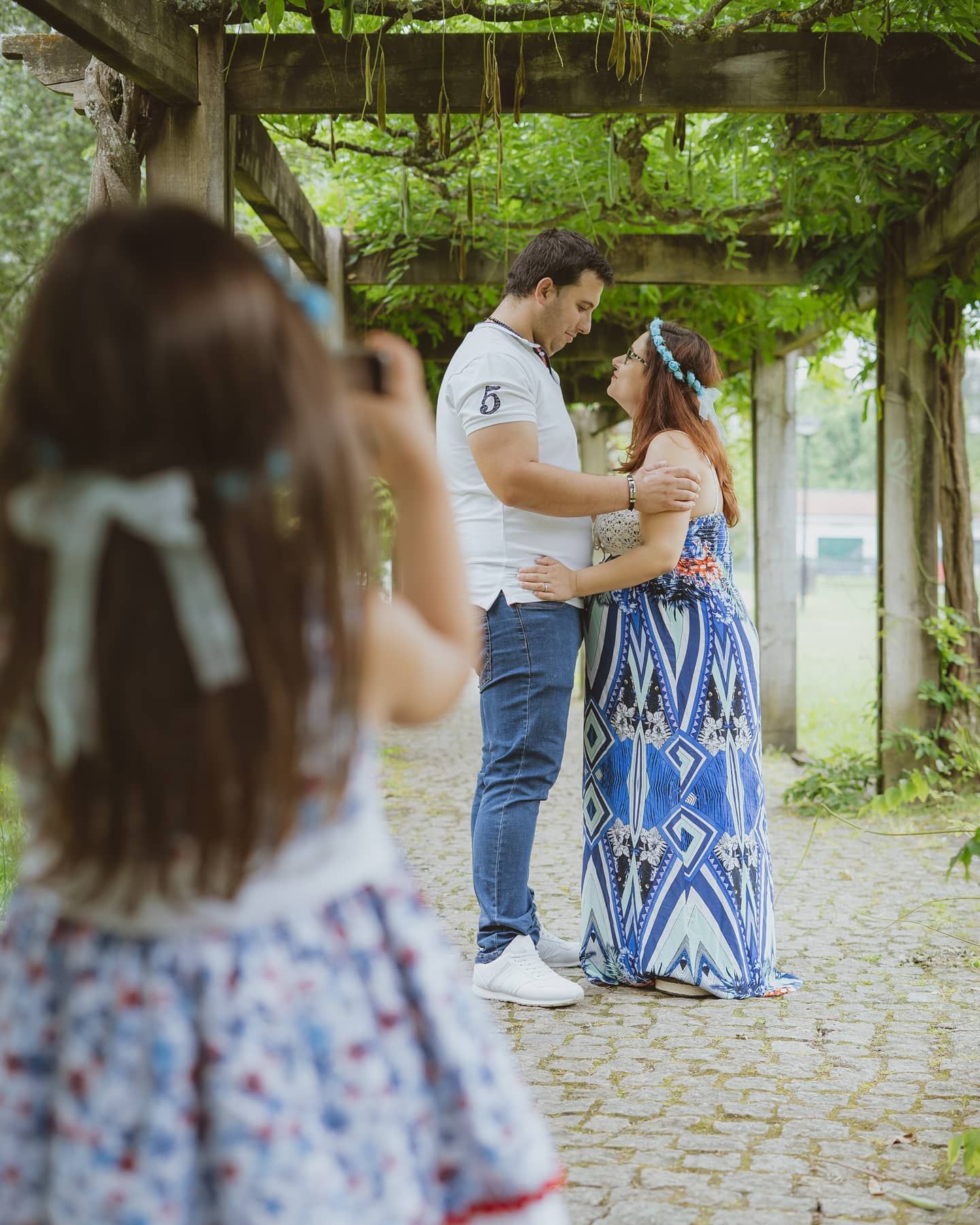 Couple embracing under a wooden pergola while a young girl photographs them in a lush garden setting.