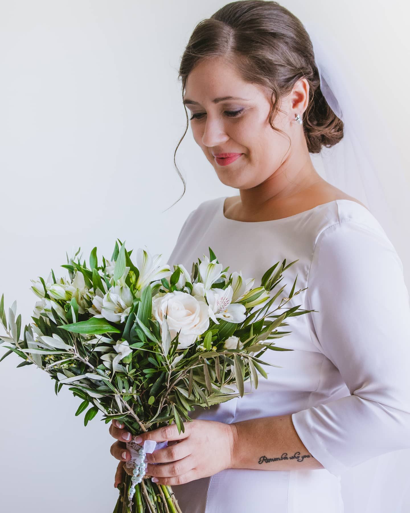 Elegant bride in white long-sleeve gown holds a bouquet of white roses and greenery.