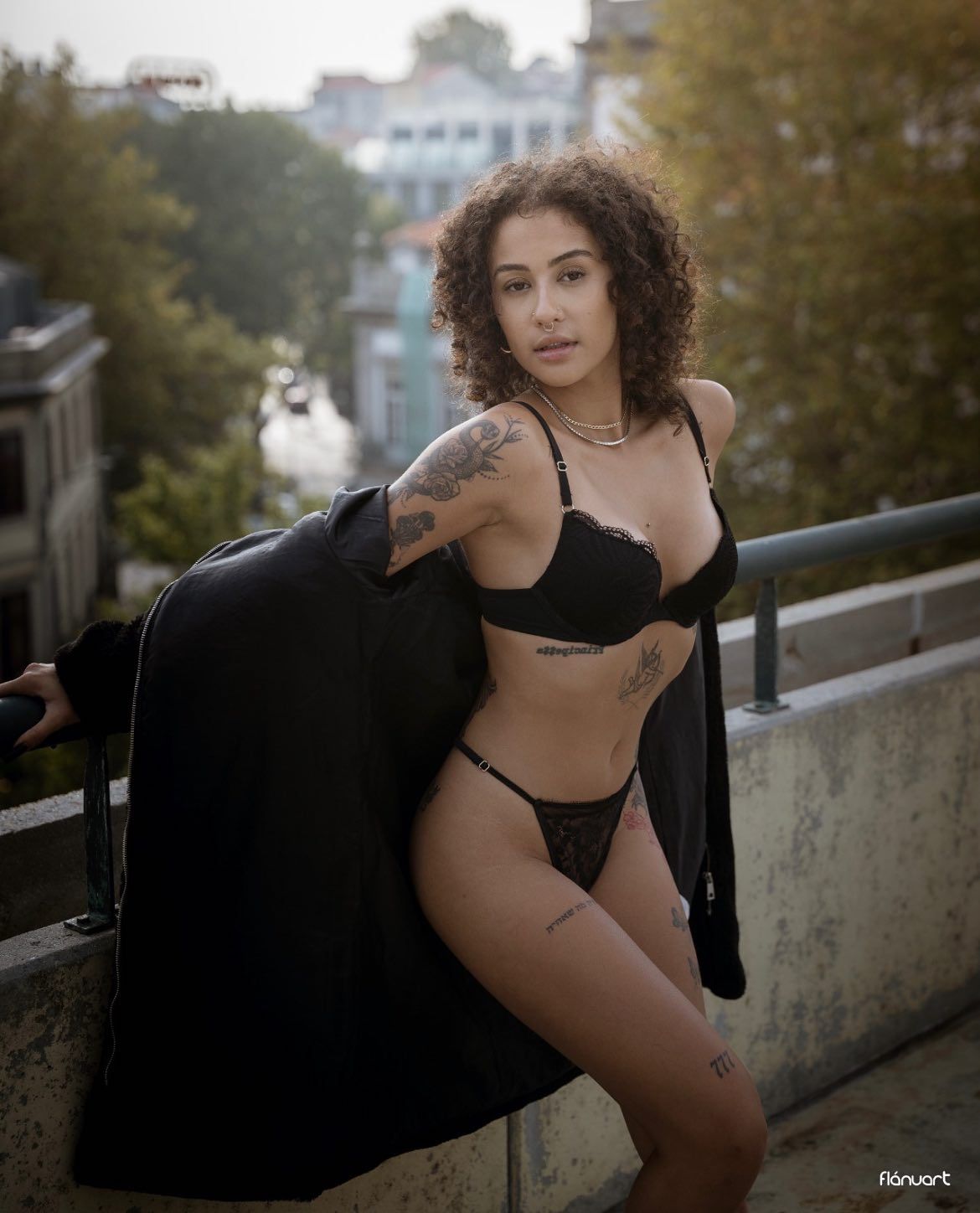 Tattooed woman in black lingerie poses on urban balcony wearing an open coat.