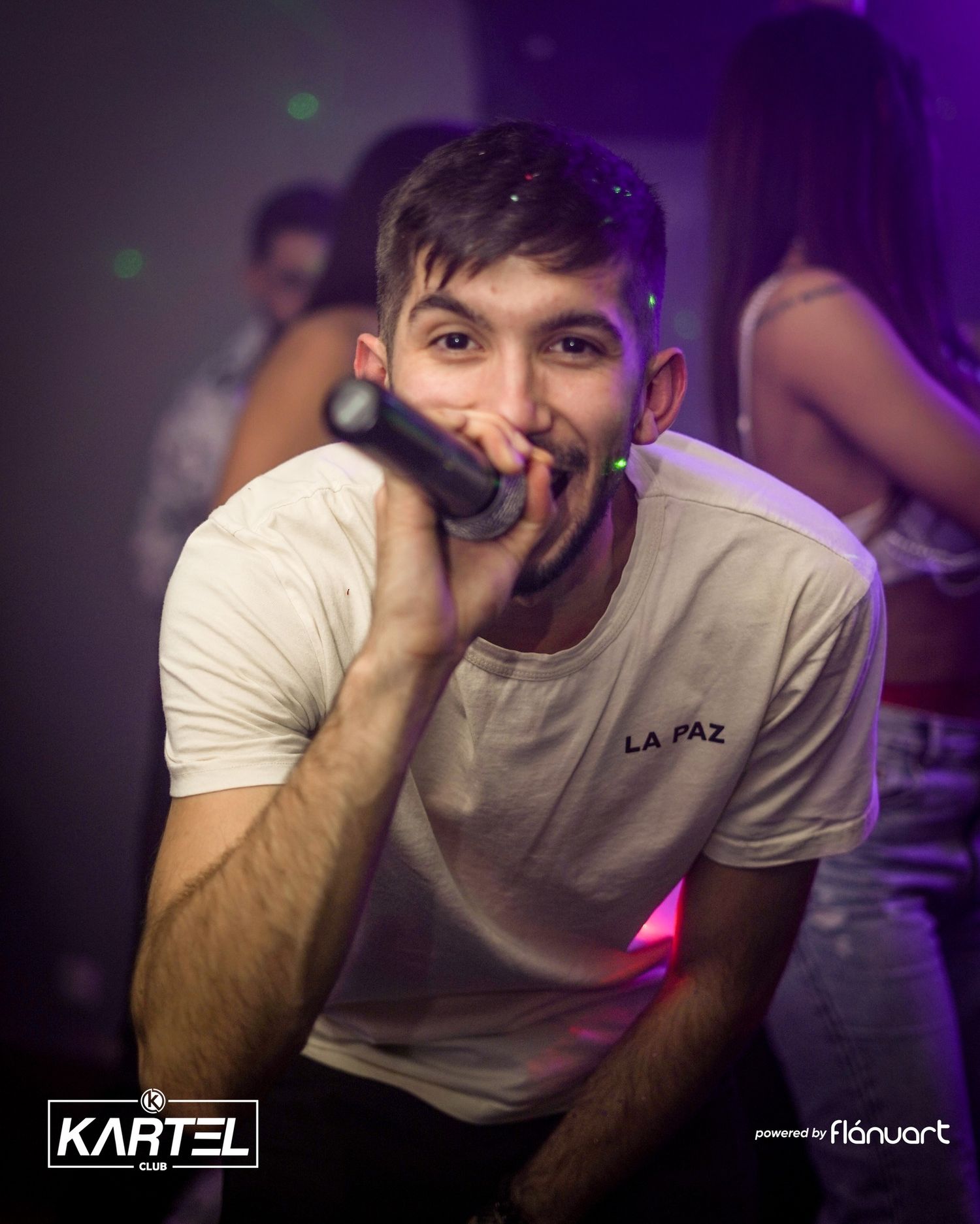 Young man in white La Paz t-shirt holding microphone and smiling at Kartel Club.