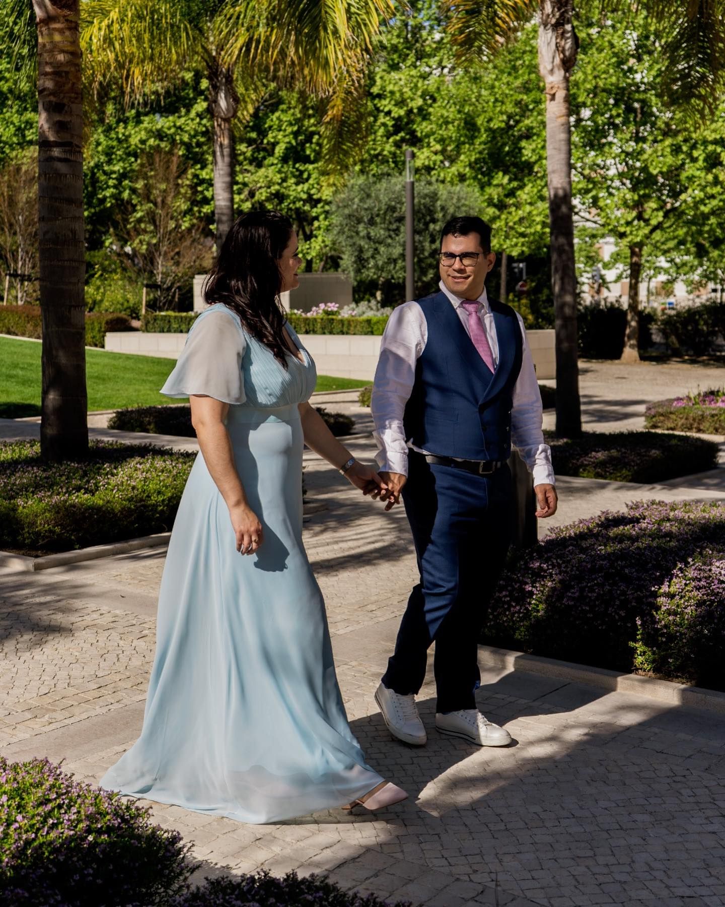 Couple walks hand in hand through a garden, woman in light blue gown, man in navy suit.