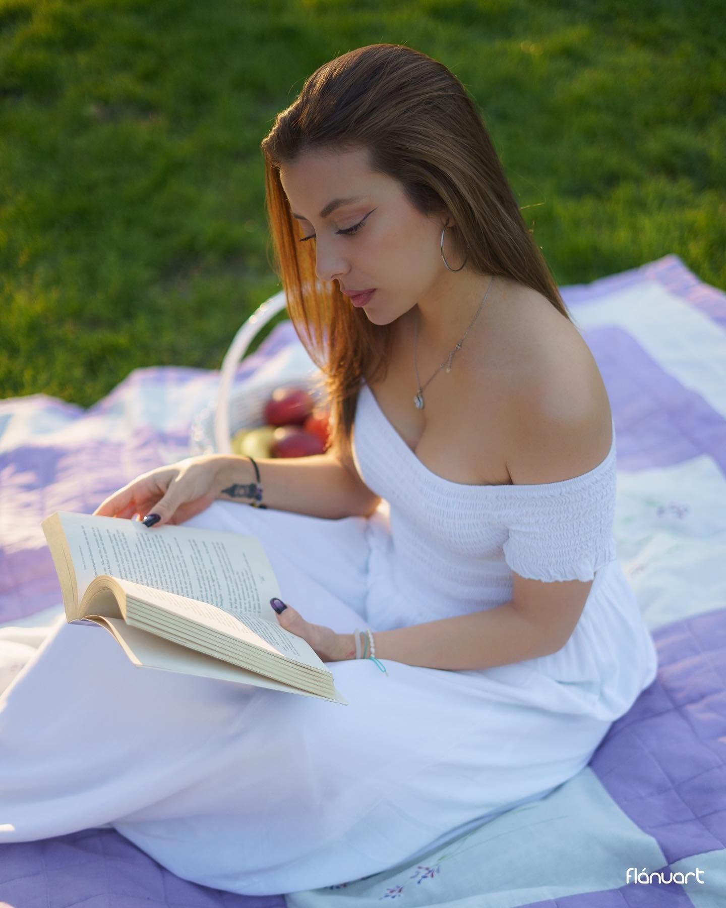Young woman in a white off-shoulder dress reading a book on a blanket in a green park.