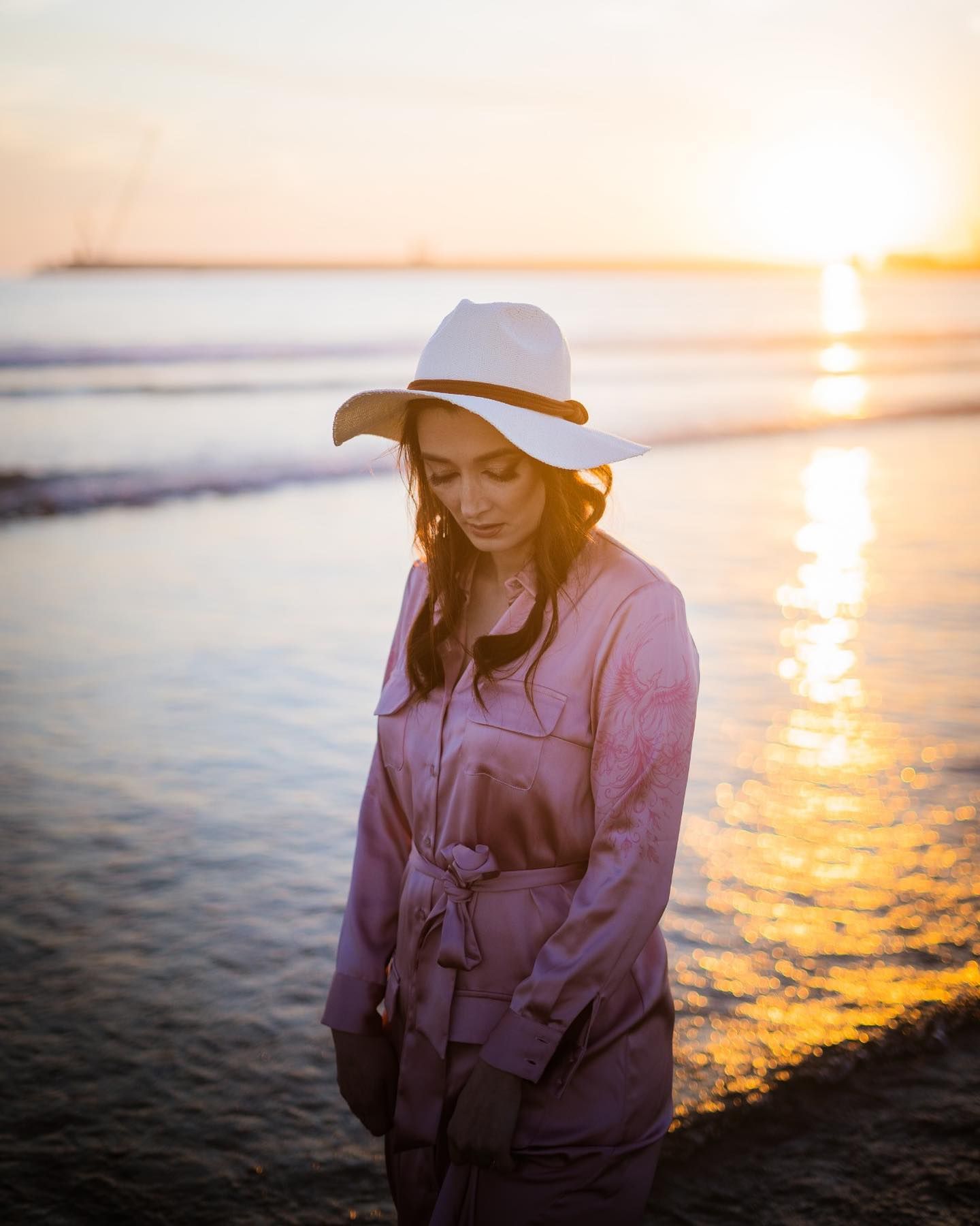 Woman in purple jacket and white hat standing on beach at golden sunset.