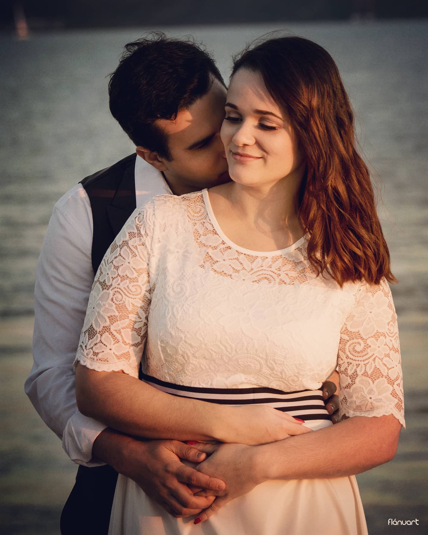 Couple sharing a romantic moment by the water at sunset, man kissing womans cheek in white lace dress.