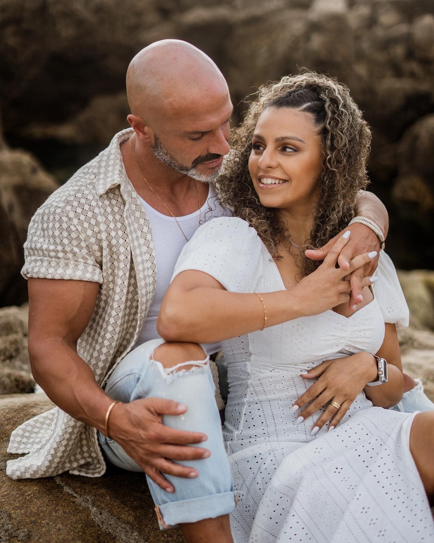 Bald man with beard and curly-haired woman in white dress sitting on rocks, smiling at each other.