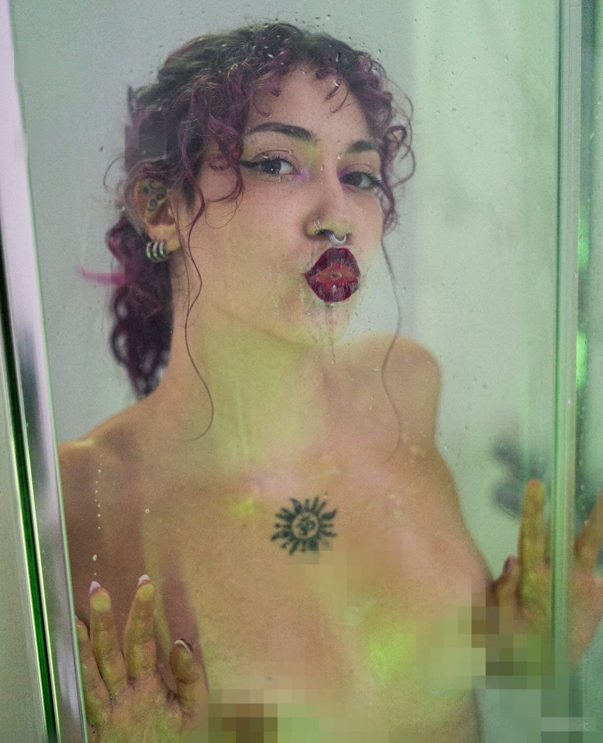 Curly-haired woman with sun tattoo pressing against glass shower door with colorful lighting.