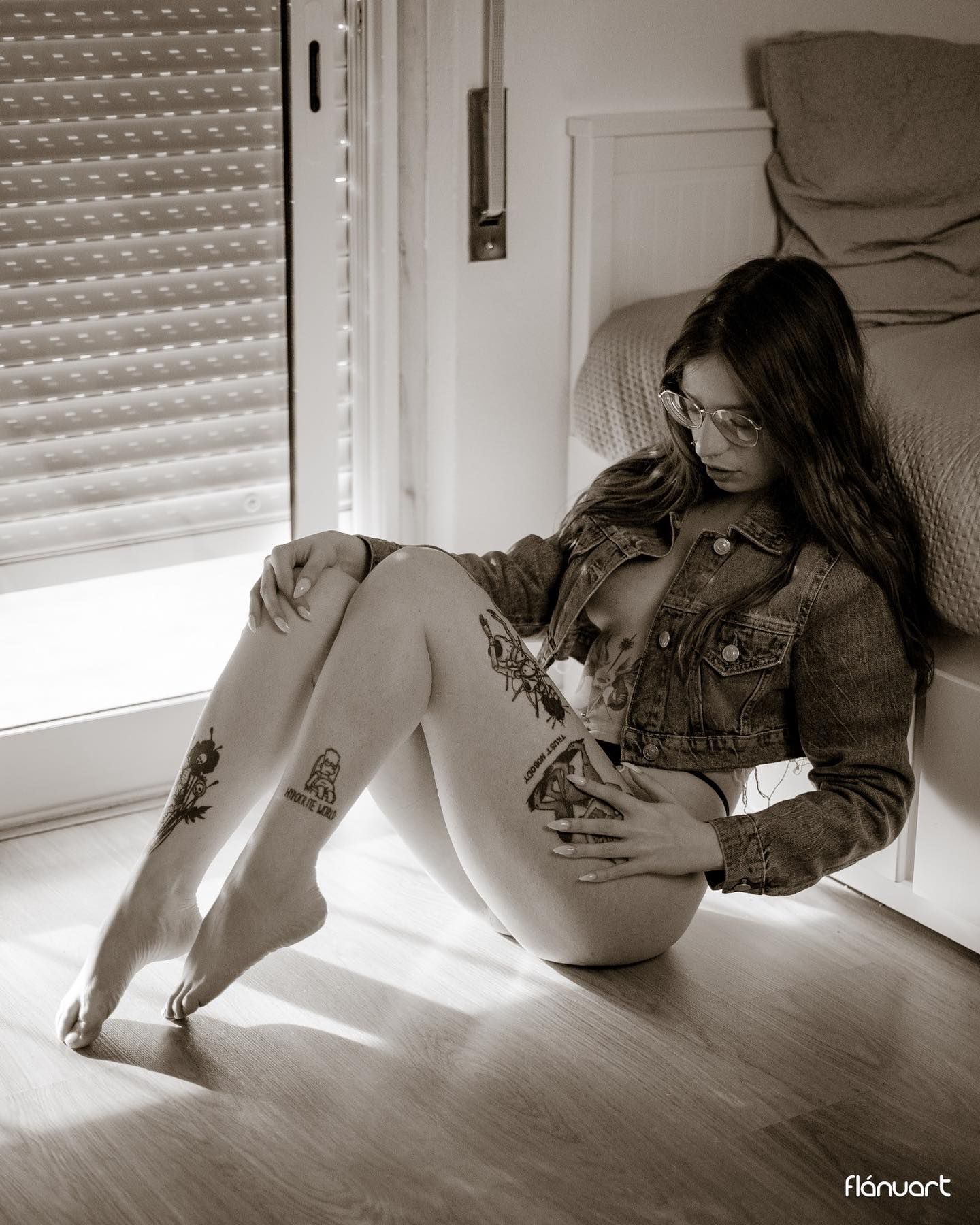 Tattooed woman in denim jacket sitting on floor with bare legs in black and white photo.