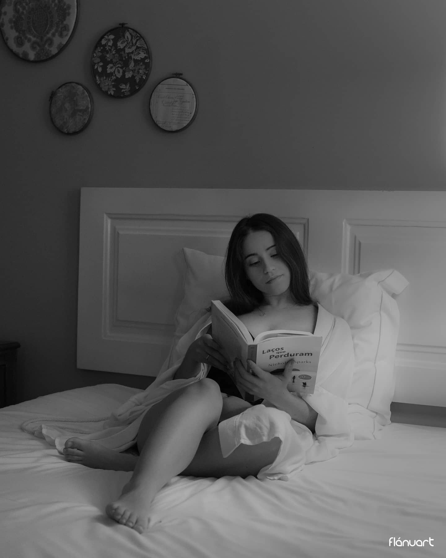 Black and white photo of a woman reading a book on a bed with pillows.