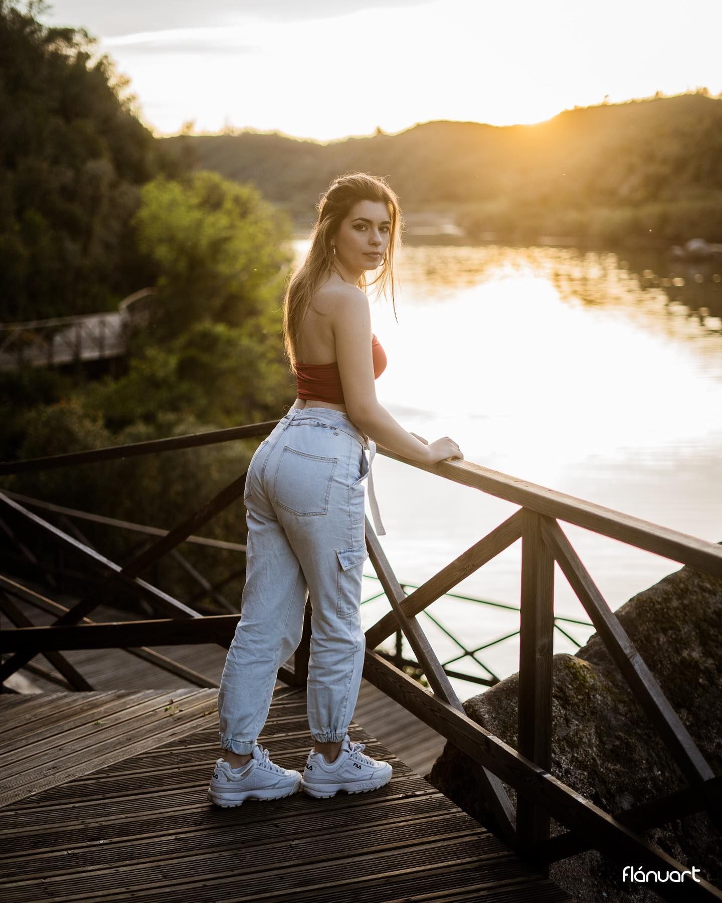 Young woman in red crop top and jeans posing on wooden bridge at golden sunset by river.