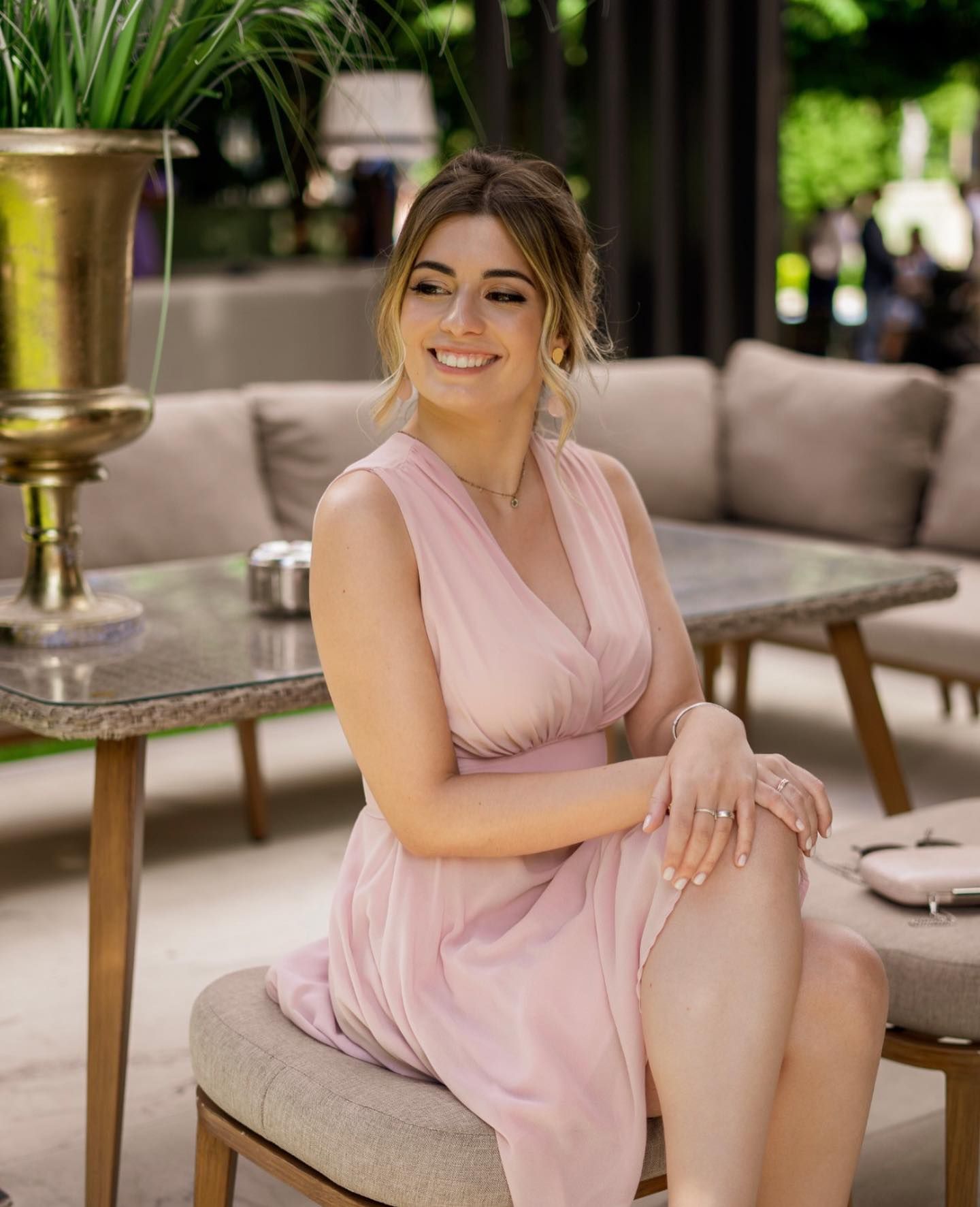 Smiling woman in pink sleeveless dress seated outdoors in elegant garden lounge setting.
