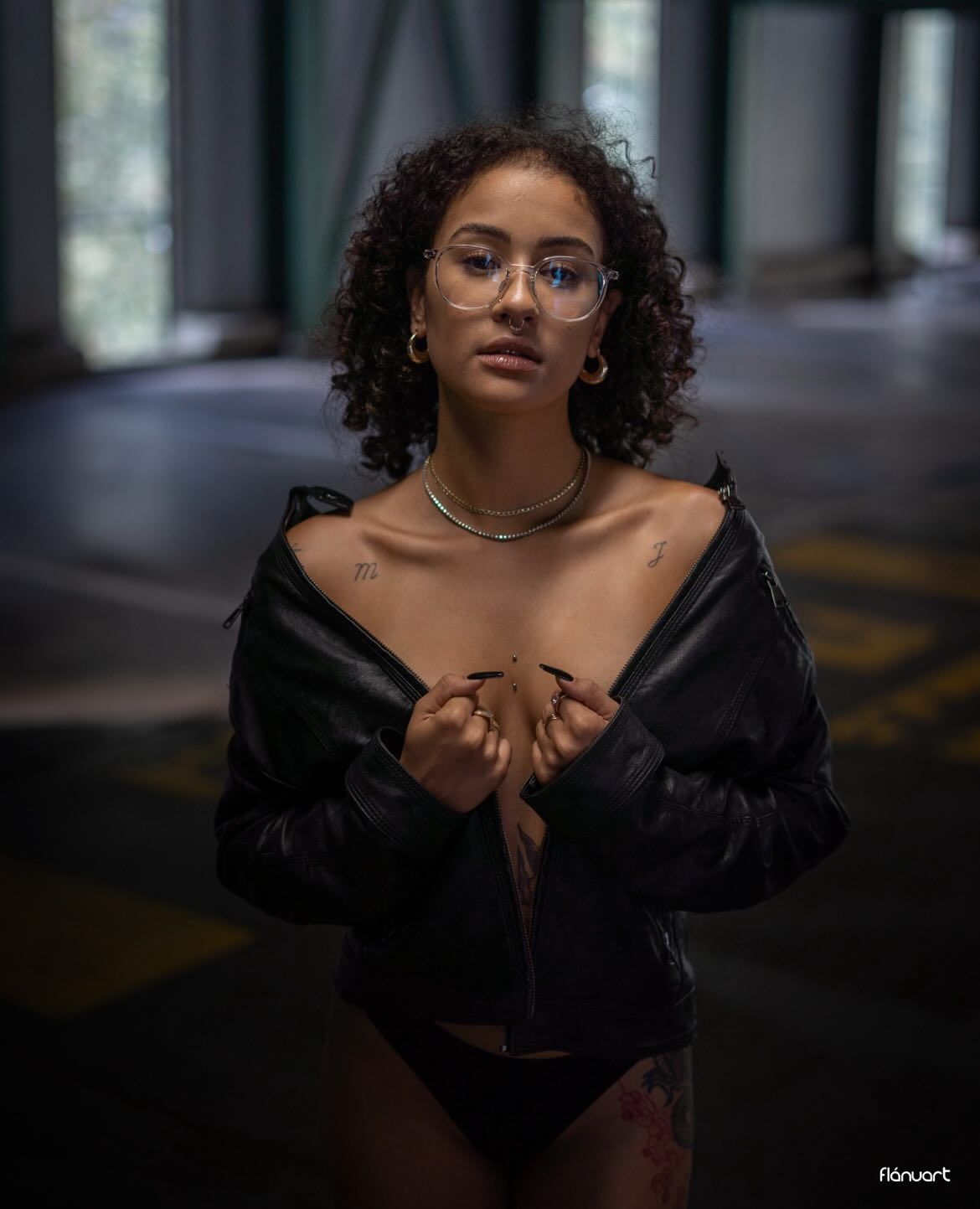 Young woman with curly hair and glasses in open black jacket posing in a dimly lit urban parking structure.