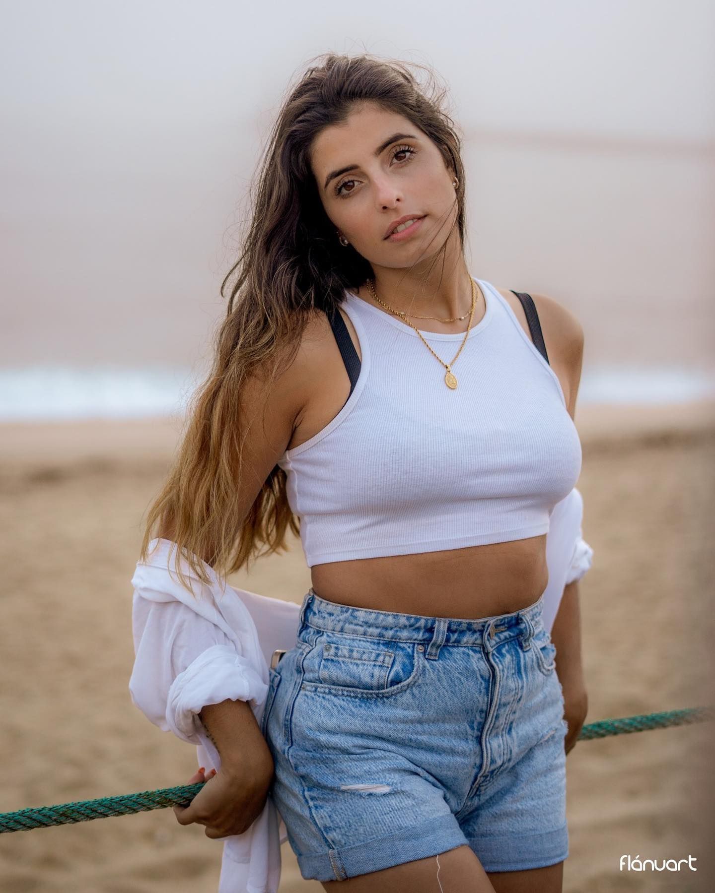 Young woman in a white crop top and denim shorts posing on a foggy beach with rope.
