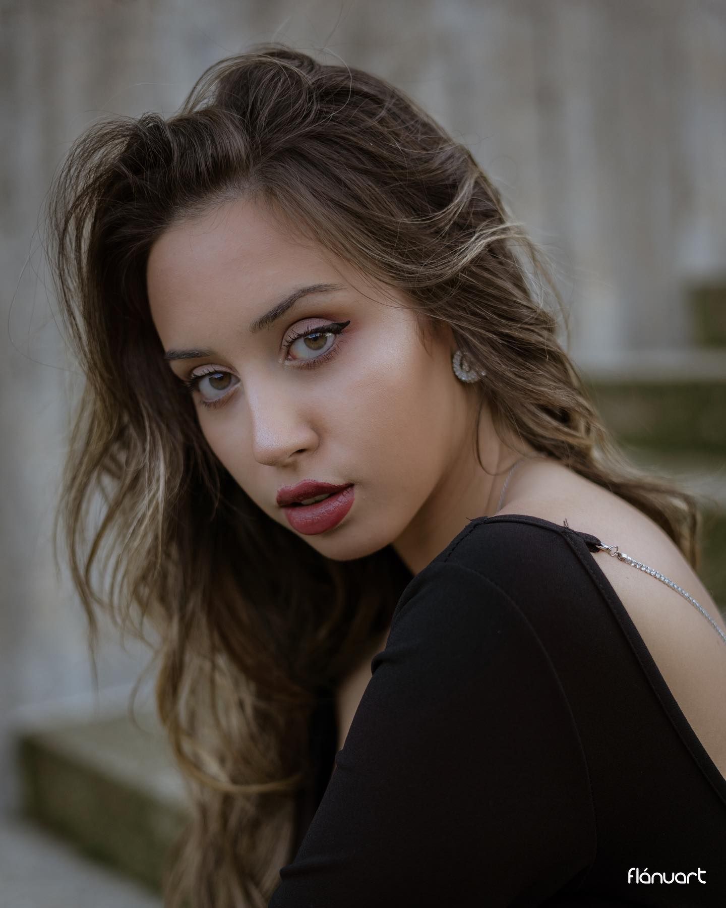 Young woman with wavy brown hair in a black top posing outdoors against a stone wall.