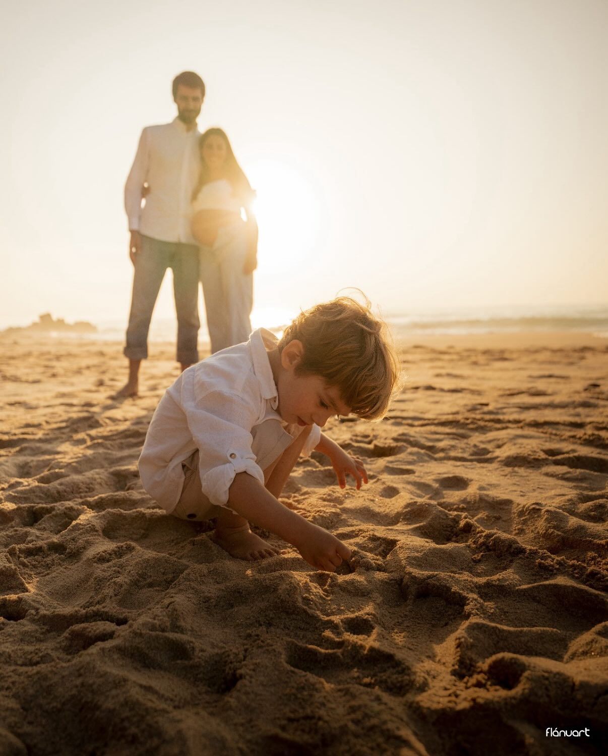 Child playing in sand on beach at golden sunset with parents standing behind.