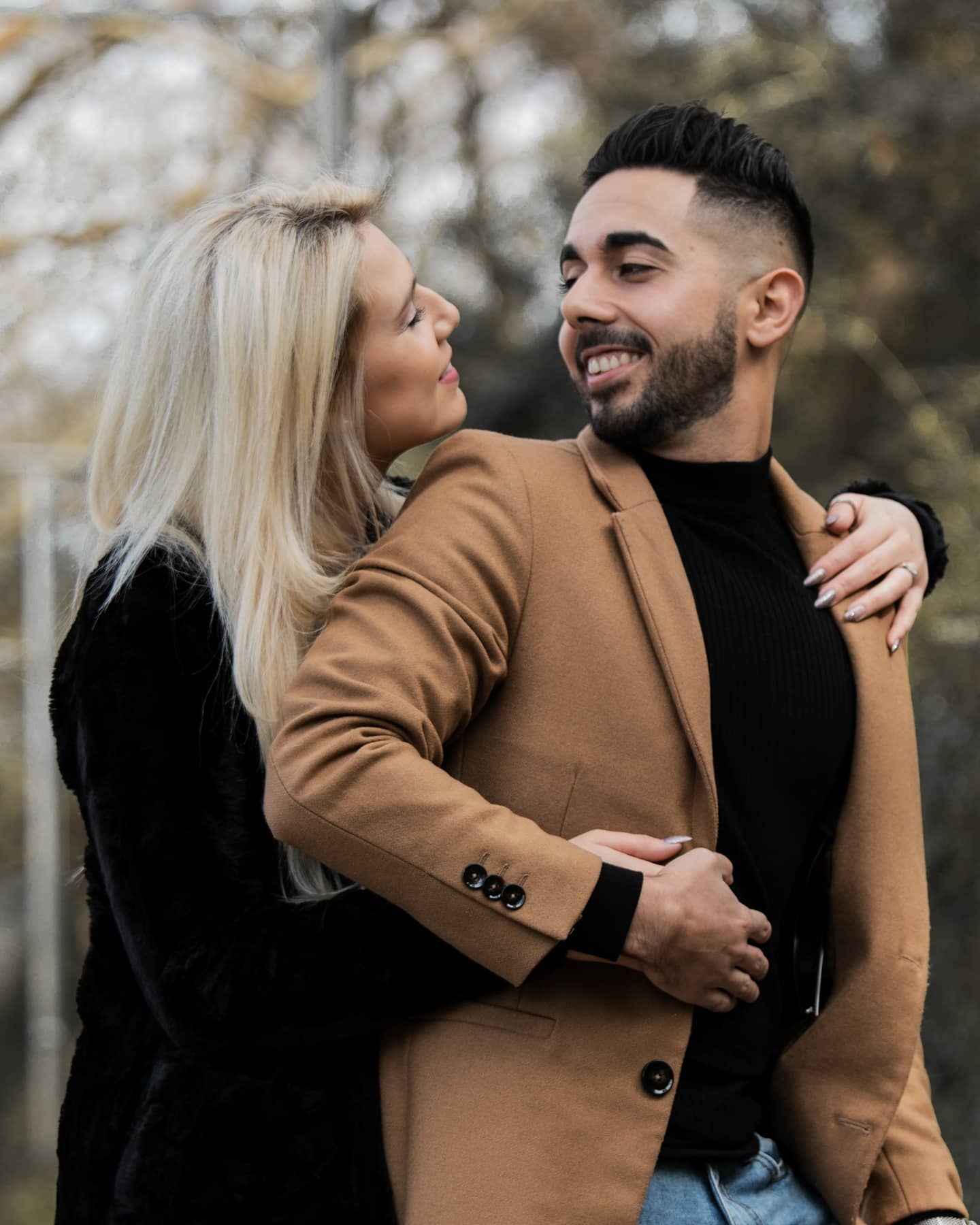 Happy couple embracing outdoors, man in camel blazer and black turtleneck, woman in black velvet.