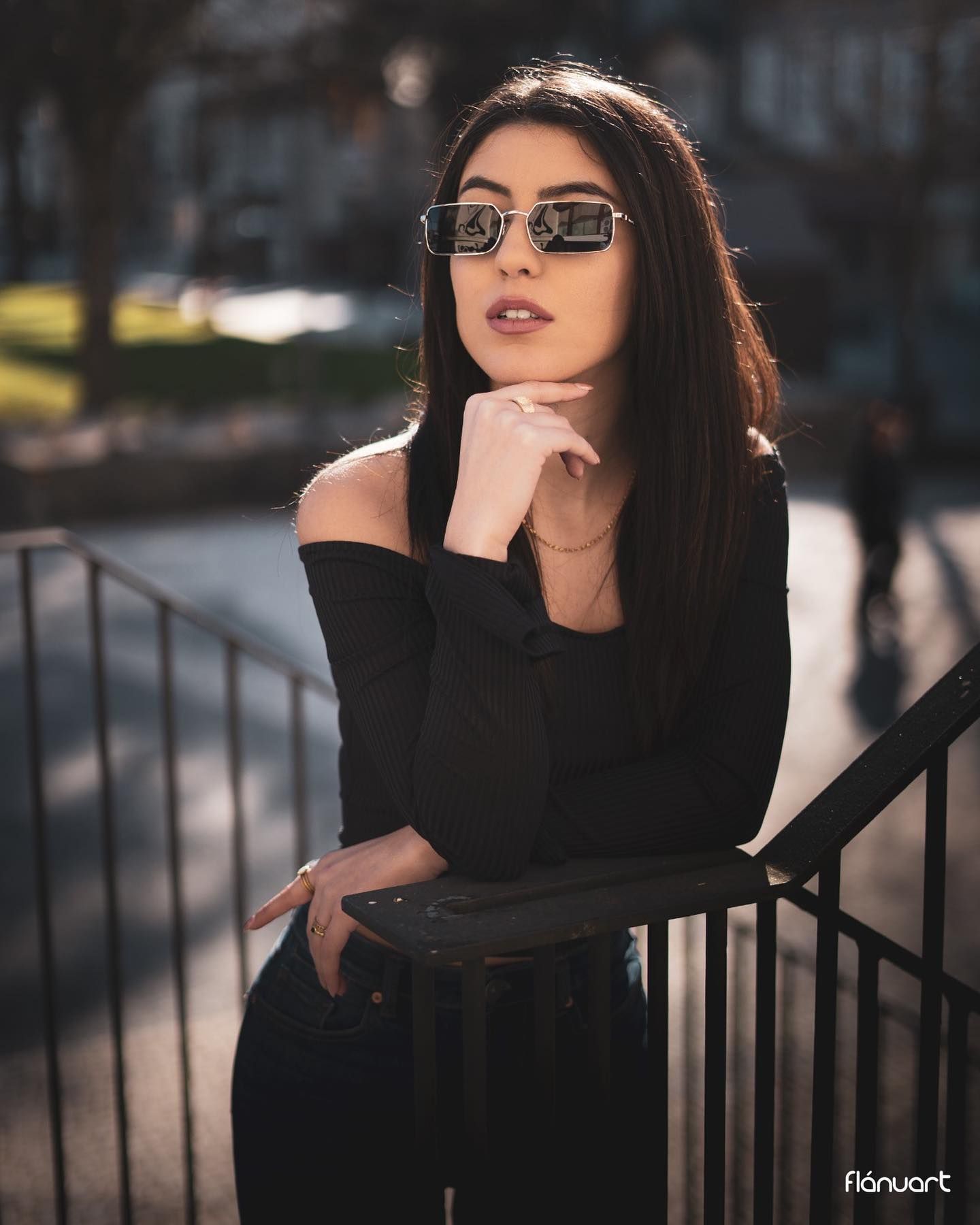 Brunette woman in black off-shoulder top and sunglasses posing on outdoor stairs in sunlight.