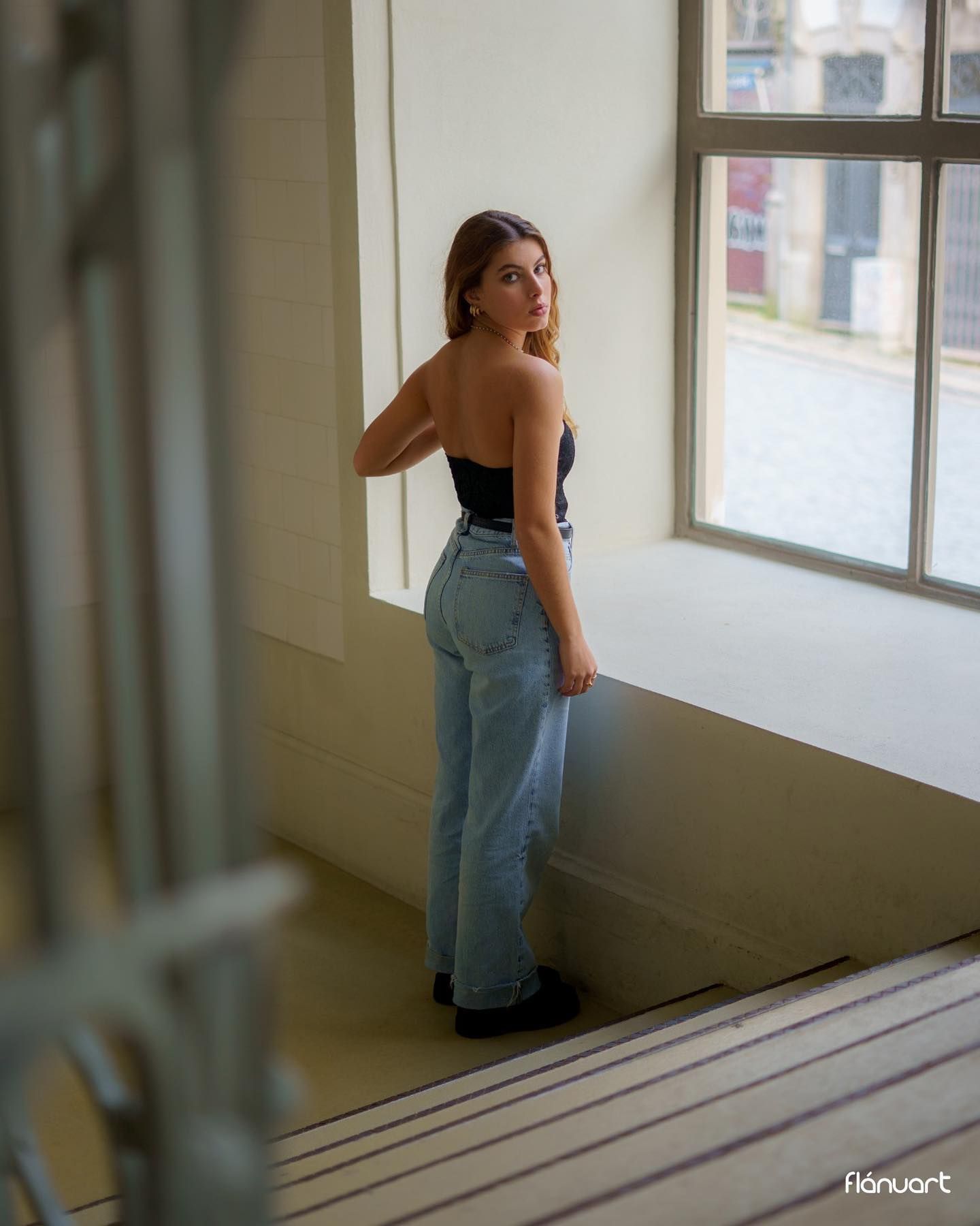 Woman in black strapless top and wide-leg jeans posing by window in bright indoor setting.