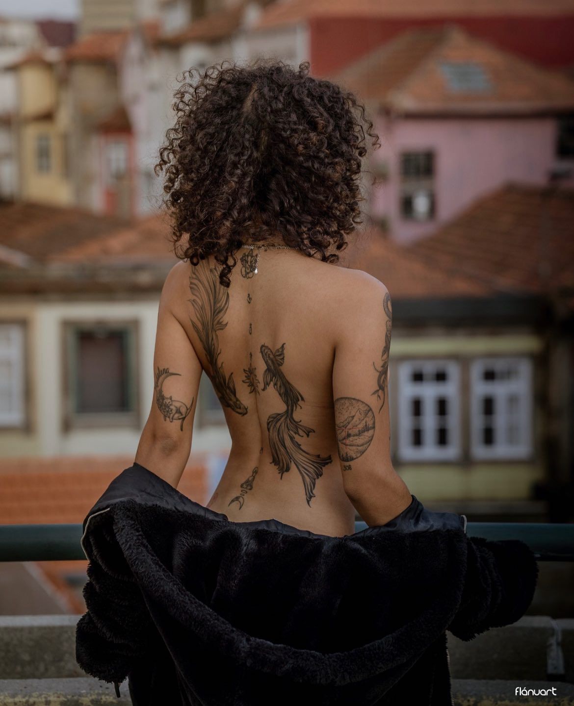 Woman with curly hair showing intricate back tattoos wearing an open black jacket outdoors.