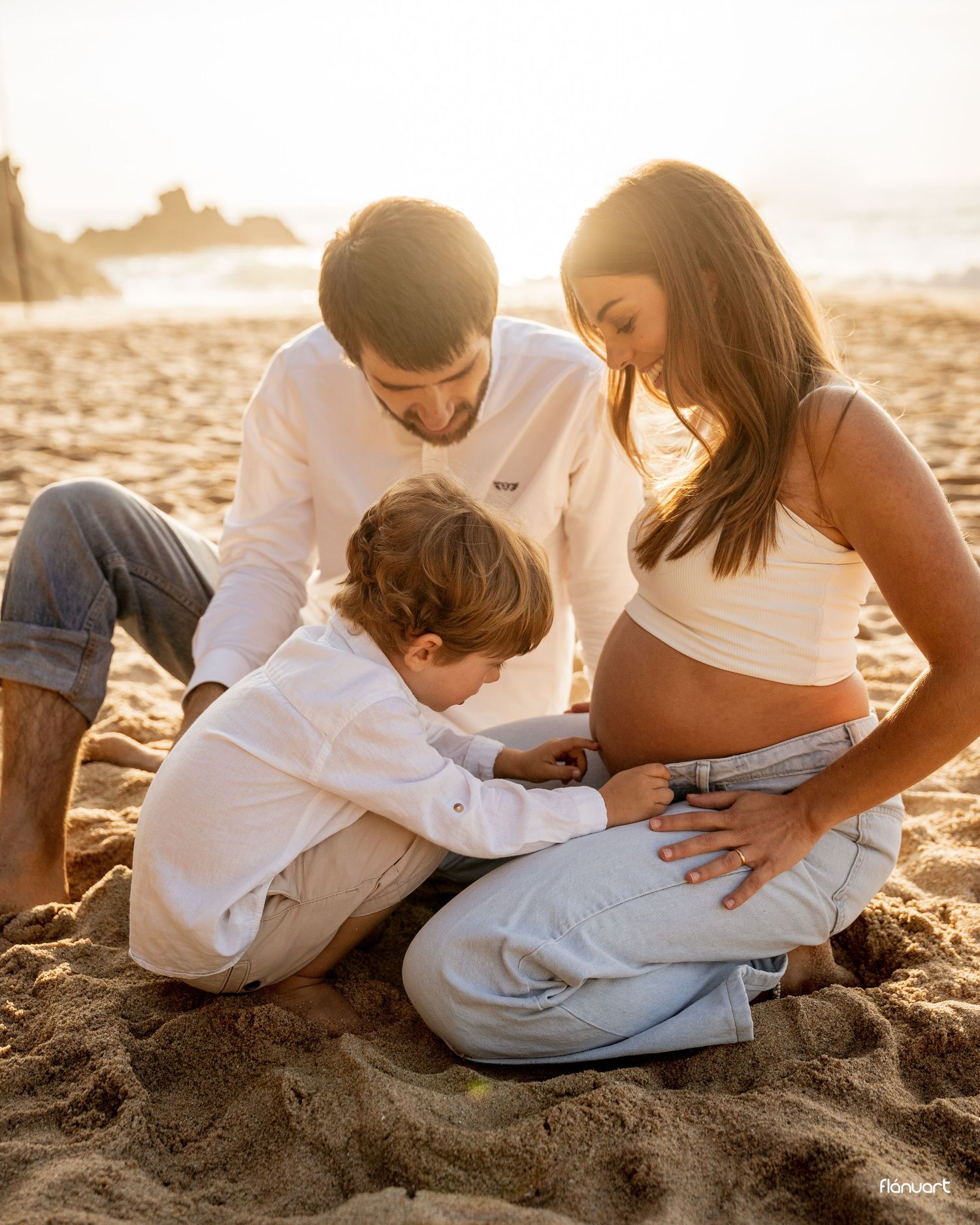 Expecting family at beach sunset, young boy touching mothers pregnant belly tenderly.