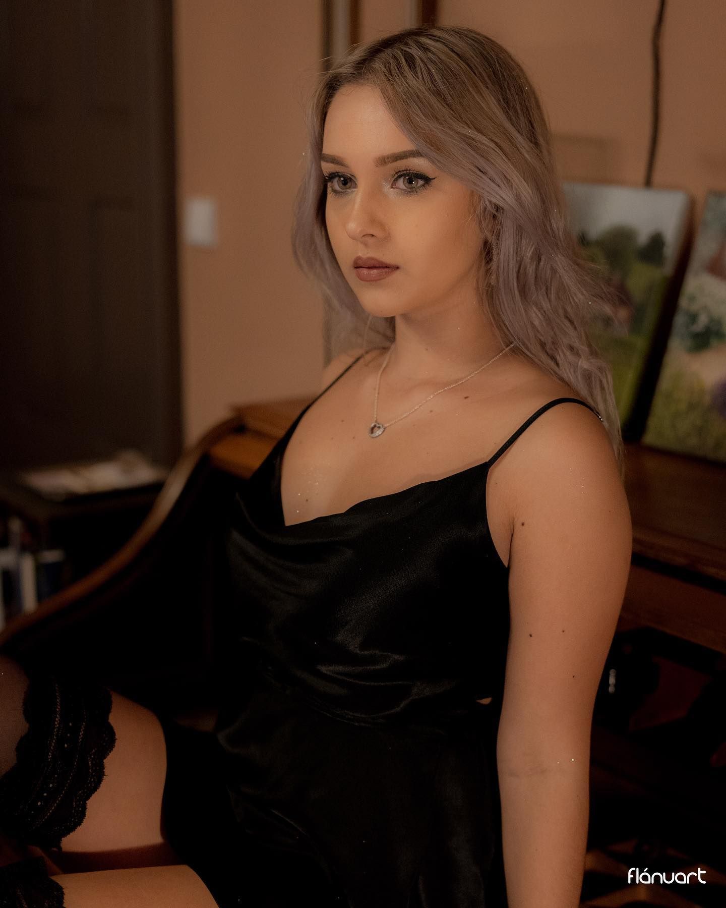 Young woman with silver hair wearing black satin slip dress, seated in artistic indoor setting.