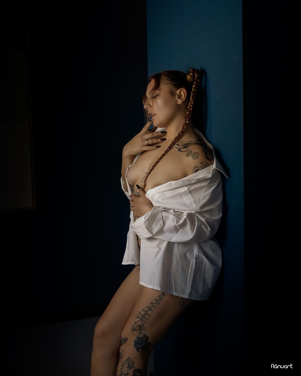 Tattooed woman in open white shirt posing against blue wall in dramatic lighting.