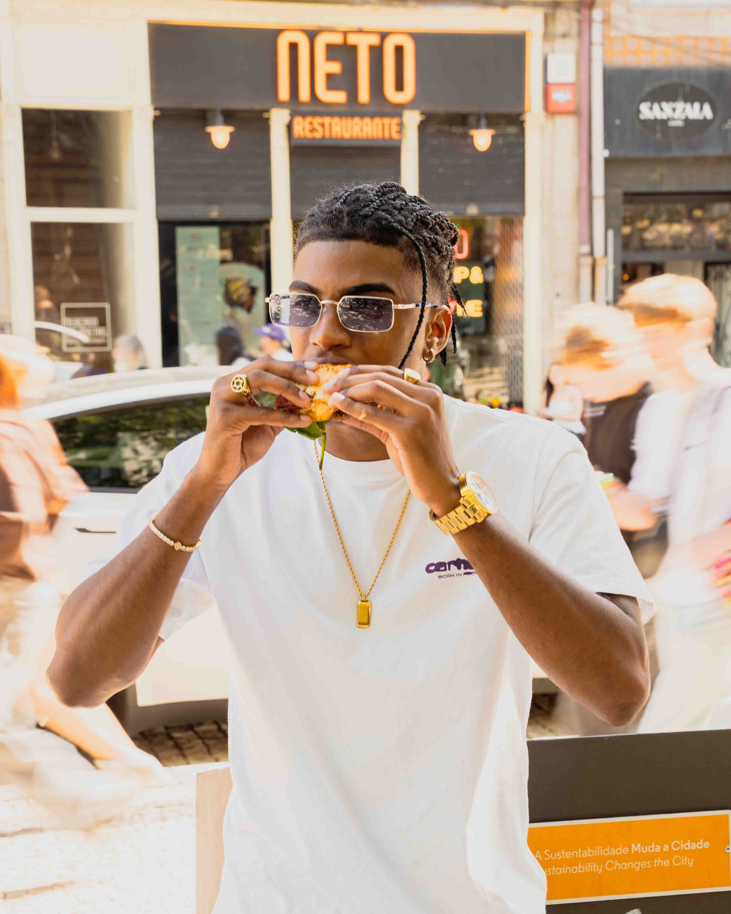 Young man in white t-shirt eating a sandwich outside Neto Restaurante wearing gold jewelry.