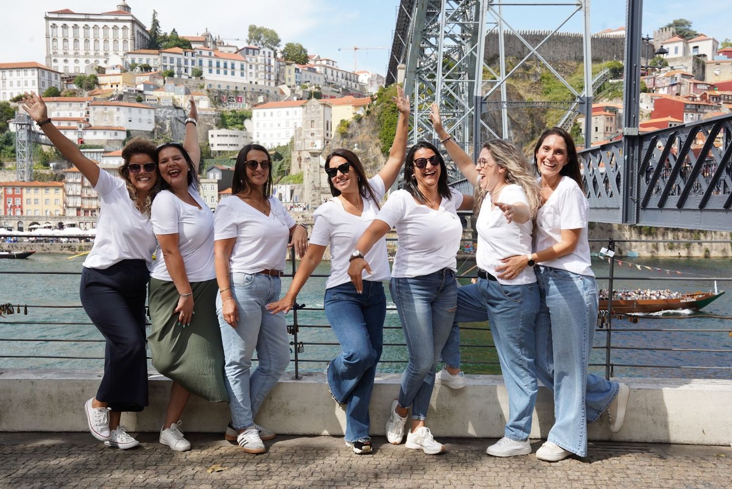Seven women in white tops and jeans posing and waving near Porto's iconic Dom Luis I bridge.